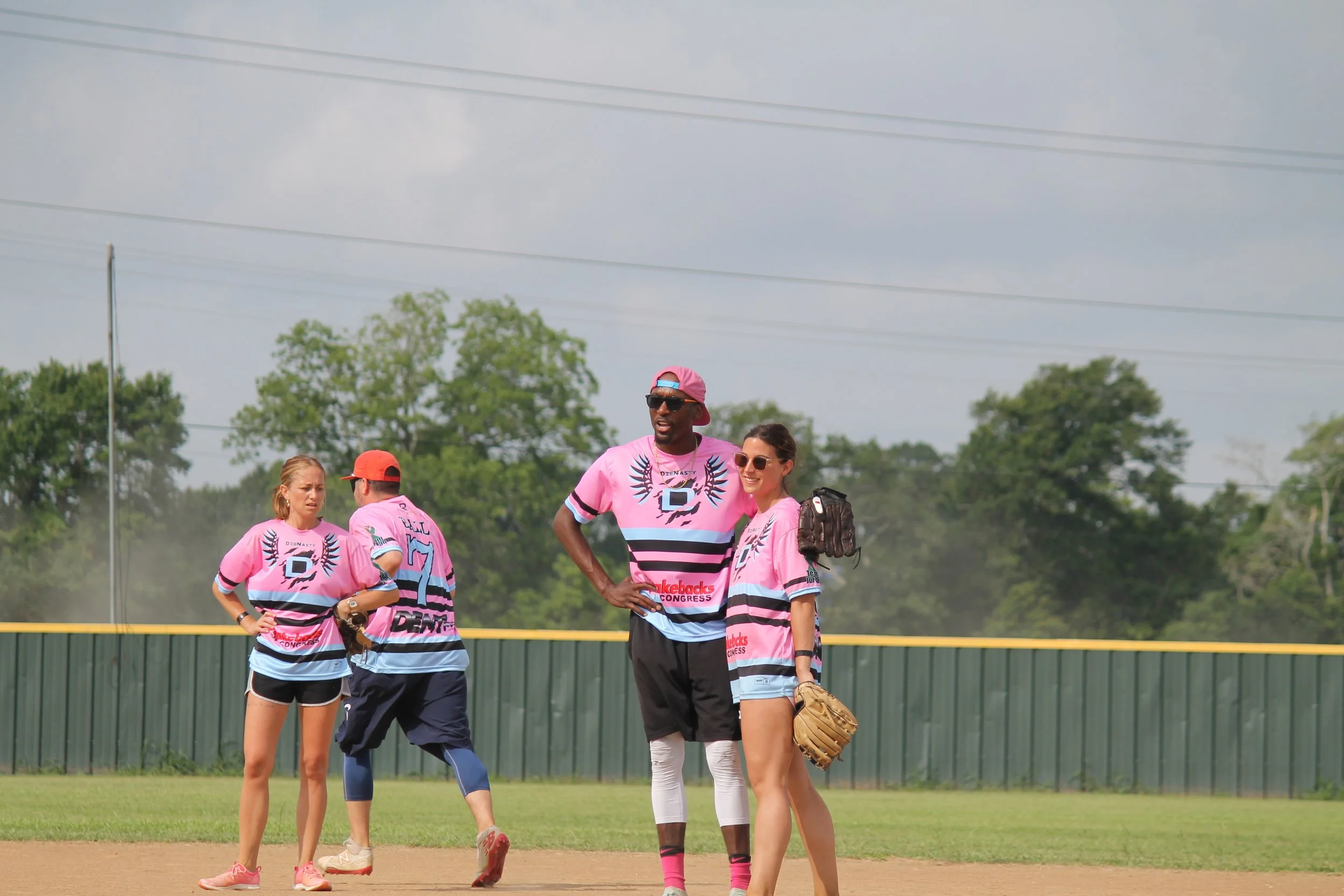 Group of people on a baseball field wearing pink jerseys, three are engaged in conversation while two are facing away, with trees and a cloudy sky in the background.