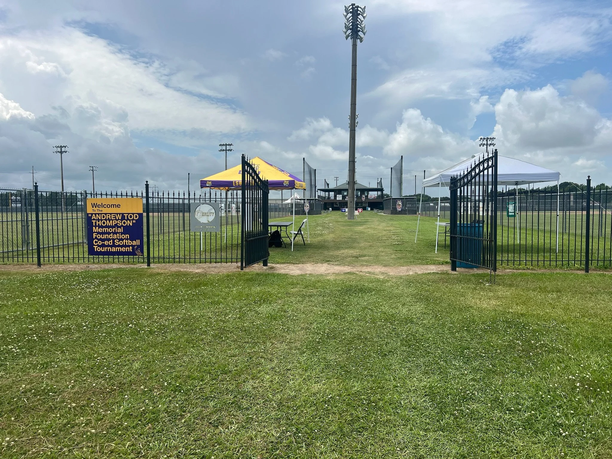 Entrance to a softball field with open gate, sign welcoming to Andrew Tod Thompson Memorial Foundation Co-ed Softball Tournament, tents and seating area on grass, and a scoreboard in the background under cloudy sky.