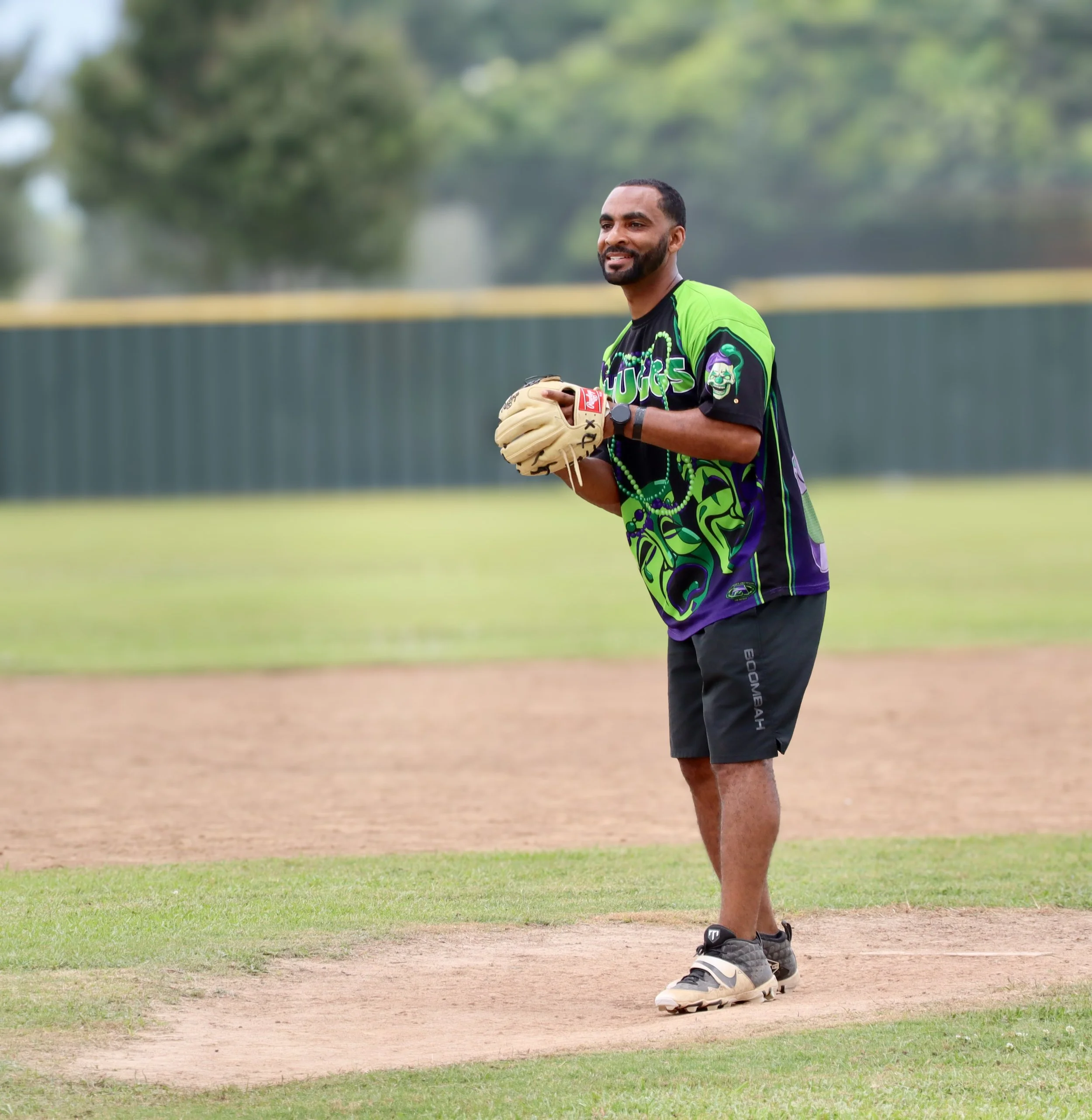 A man standing on a baseball field holding a baseball glove, dressed in sportswear, smiling, with grass and a fence in the background.