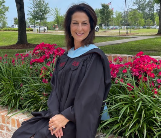 A woman in a graduation cap and gown sitting outdoors in front of pink flowers and green plants, smiling.
