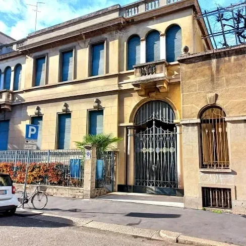 Edificio antico con pareti gialle, grandi finestre arcuate e una porta con cancellata in metallo. Sul marciapiede ci sono una bicicletta e un'auto parcheggiata, con un'area verde e un cartello di parcheggio vicino.