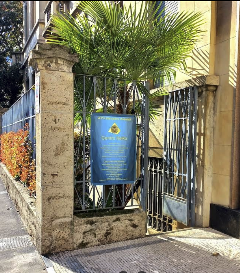 Ingresso di un edificio con cartello azzurro che indica il Centro olistico Keiko Milano andrea verga circondato da ringhiere di metallo e piante tropicali.
