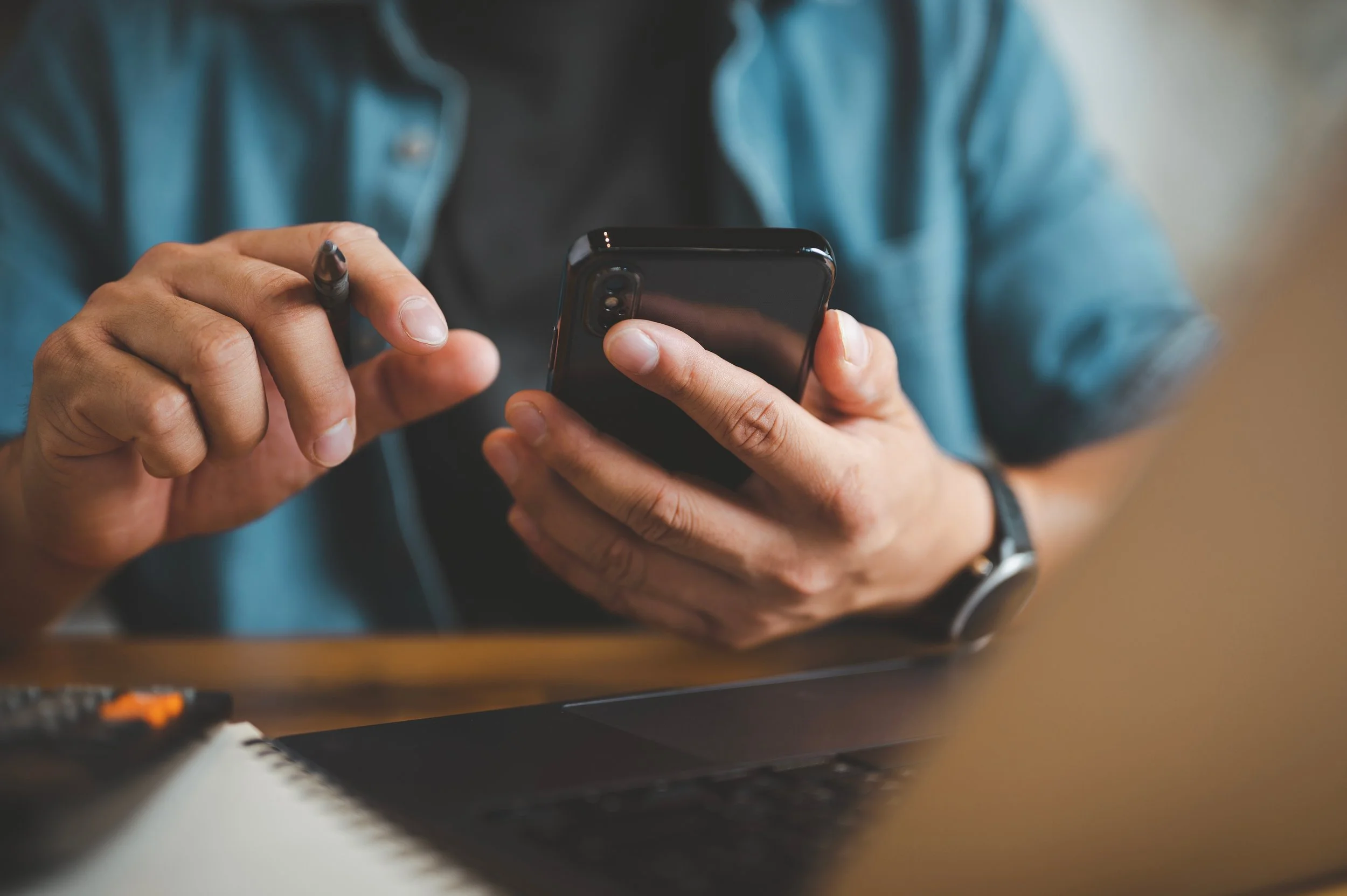 A person in a denim shirt holding a smartphone and a stylus, using the device at a desk with a laptop and notebook.