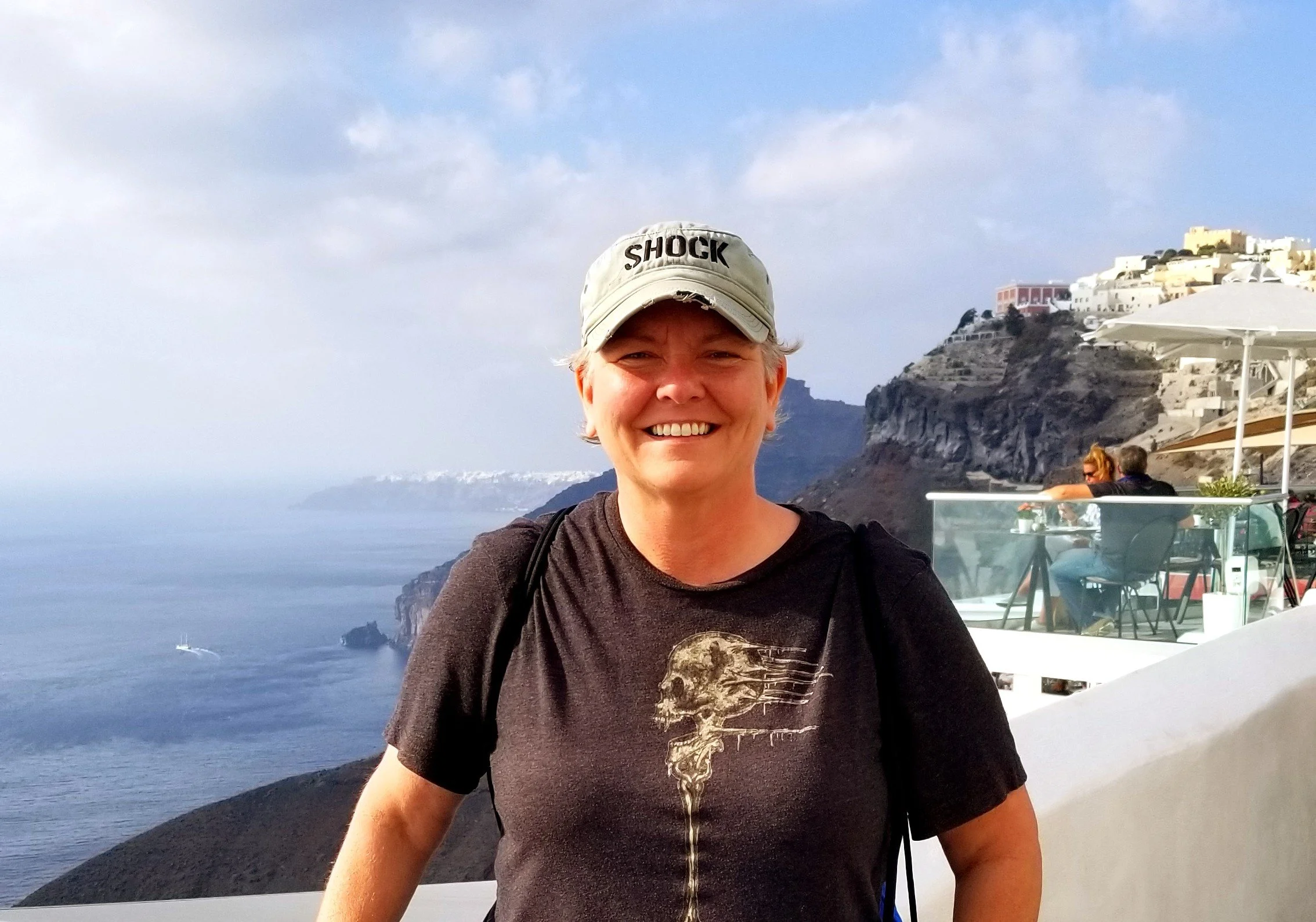 Smiling woman wearing a cap and black T-shirt with a skull design, standing in front of coastal cliffs and buildings.