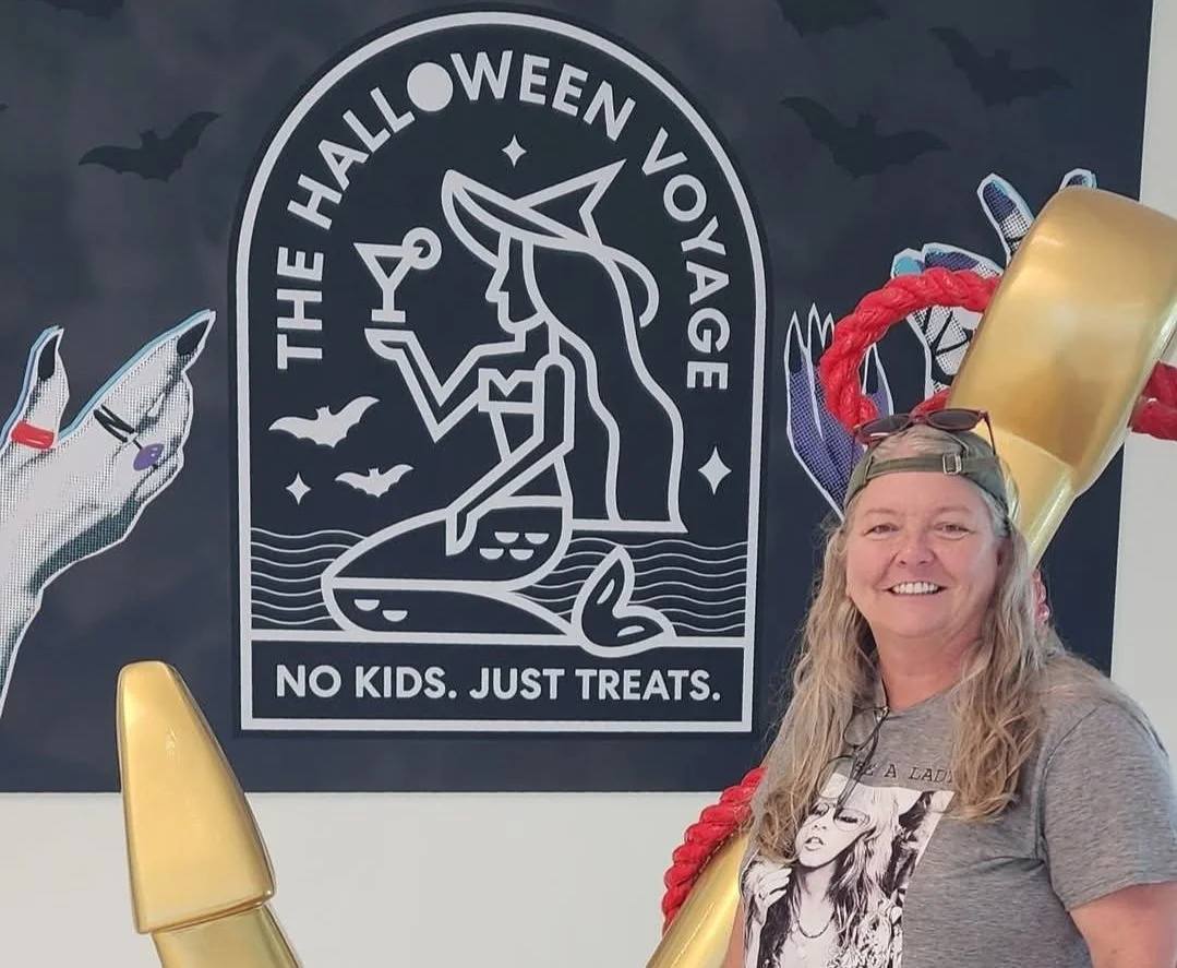 Woman smiling at Halloween event with a large sign behind her that reads 'The Halloween Voyage. No Kids. Just Treats.' and features a witch silhouette, bats, and waves.