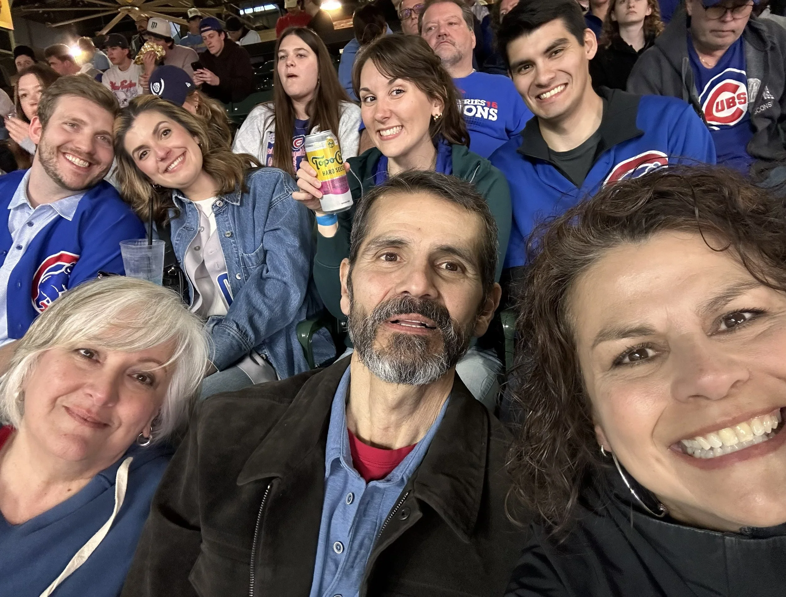 A group of people at a sports event, smiling and posing for a selfie. They are wearing Chicago Cubs gear, with a stadium background and other spectators visible.