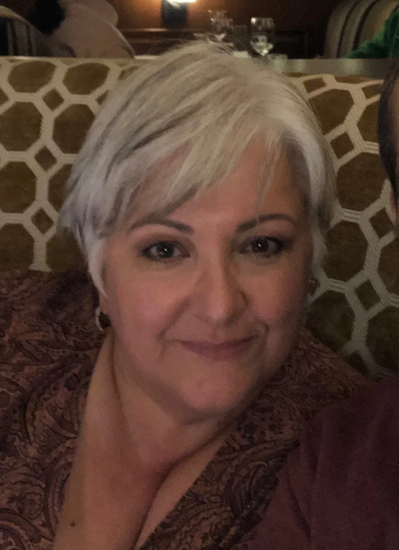 A woman with short, blonde hair smiling at the camera in a dimly lit restaurant or lounge setting.
