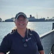 A smiling woman in a dark jacket and cap standing near a waterfront with industrial buildings and ships in the background.