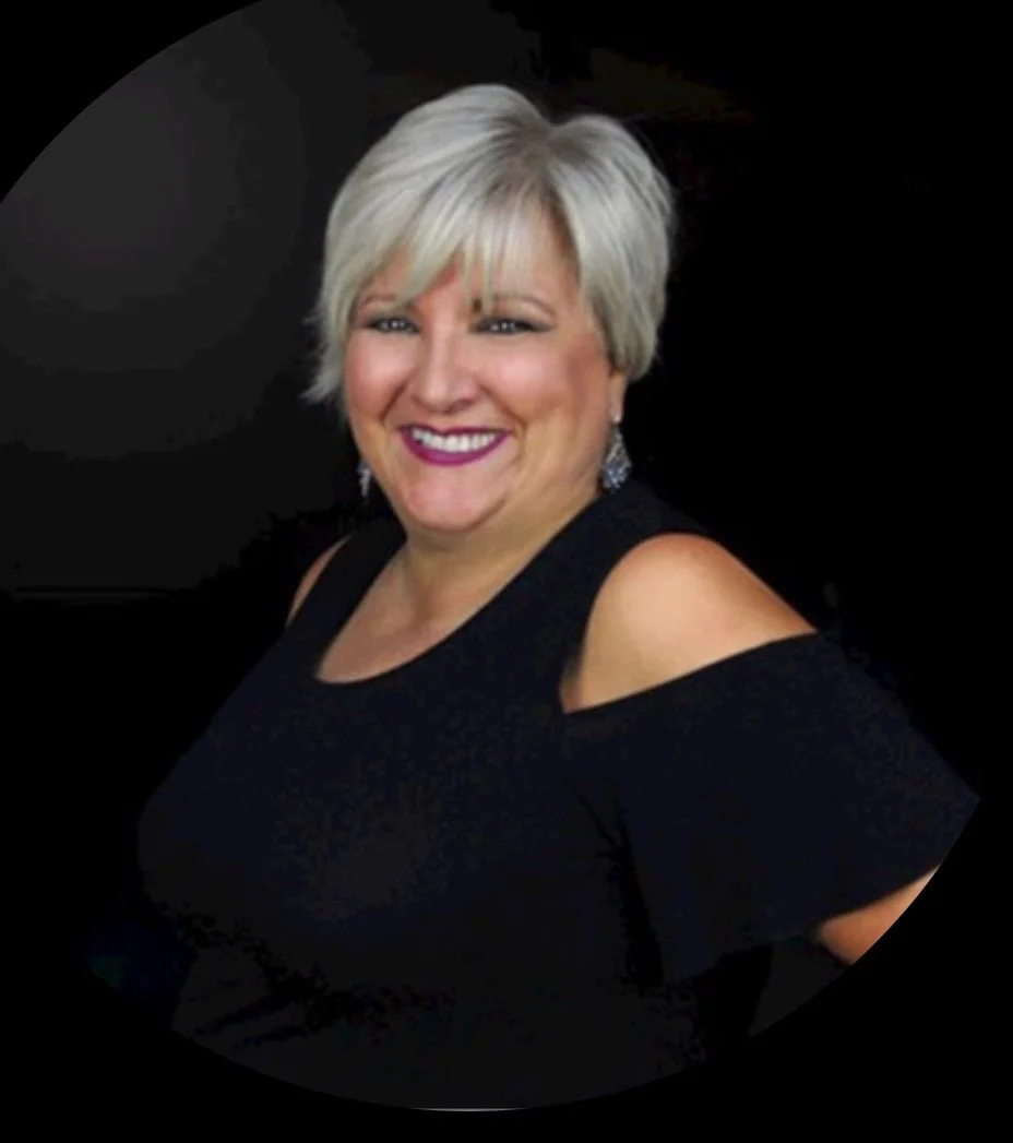 A smiling woman with short, gray hair wearing a black dress with cold shoulder sleeves and dangling earrings.