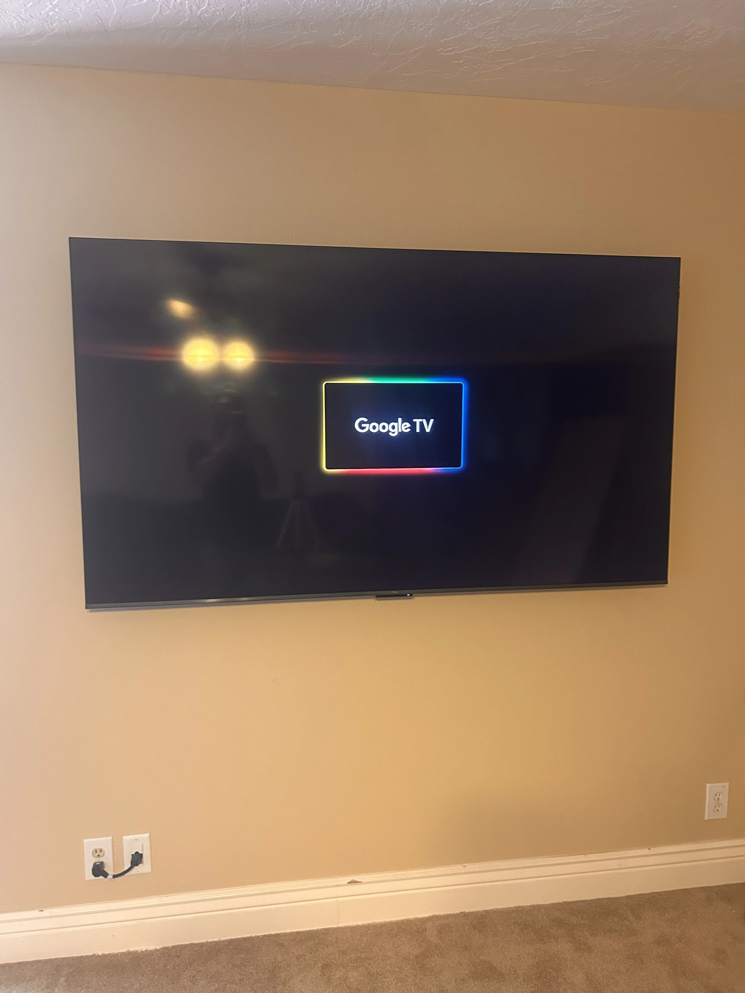 A wall-mounted flat-screen TV with the Google TV startup screen, displaying a multicolored border around the text 'Google TV'. The room has beige walls and carpeted flooring, with visible electrical outlets beneath the TV.