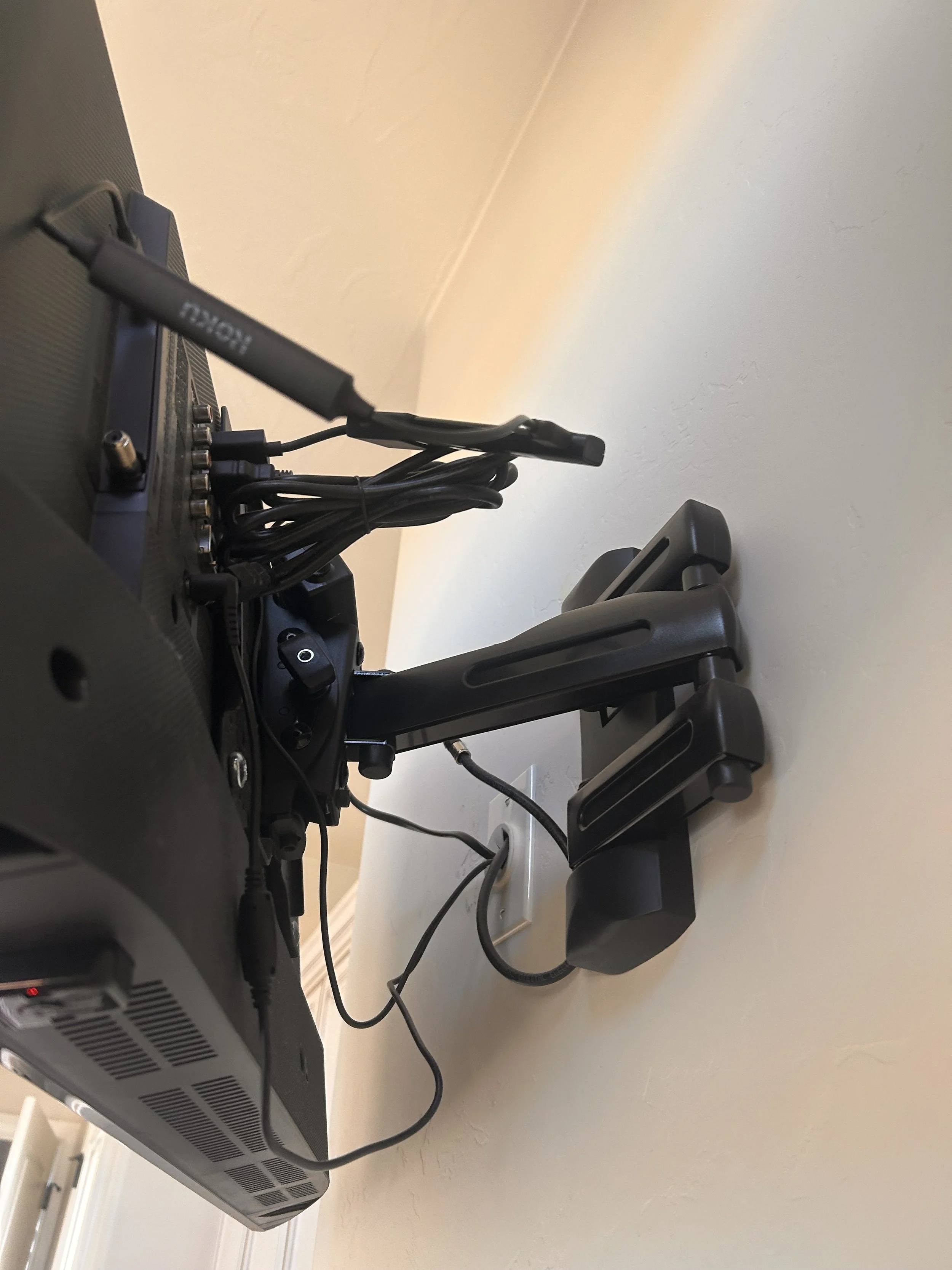 A black electronic device with various cables connected to it, mounted on a swivel arm attached to a desk or table, with a white wall in the background.