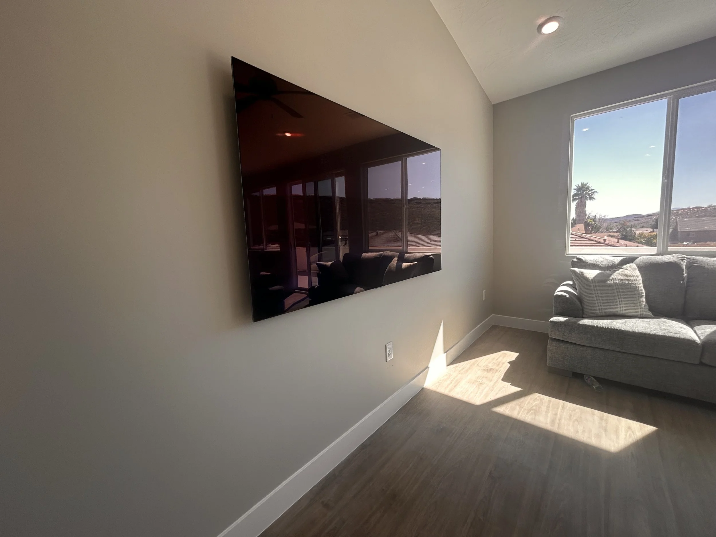 Living room with a wall-mounted flat-screen TV, a large window with a view of rooftops and a palm tree outside, and a gray sofa with pillows.