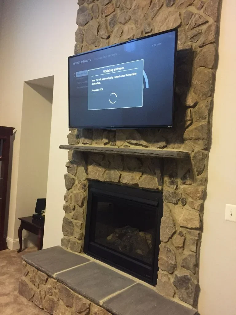 A flat-screen TV mounted above a stone fireplace with a wooden mantel, displaying a software update message, in a living room with beige walls and carpeted floor.