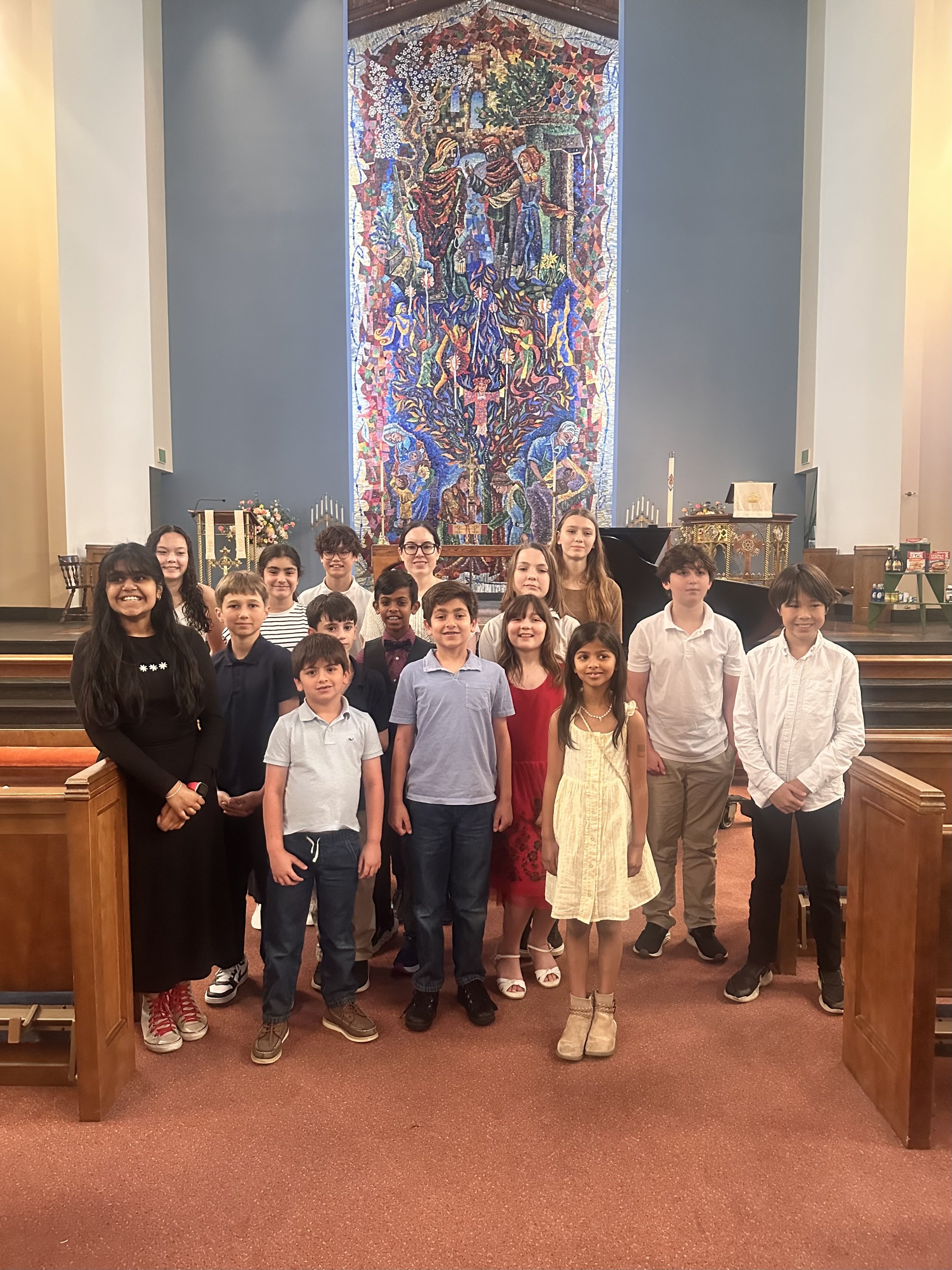 Group of children and teenagers standing inside a church, in front of a large colorful tapestry depicting a biblical scene, with an altar and candles in the background.