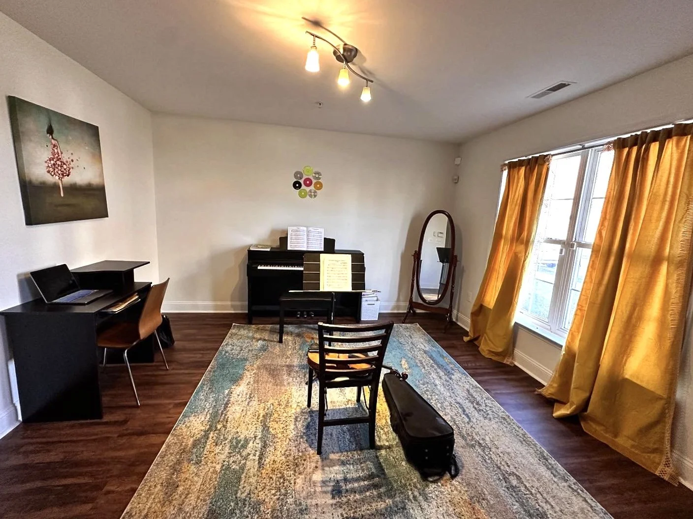 A living room with a wood floor, yellow curtains, and white walls. A black piano with sheet music stands near the wall. A mirror stands on a wooden frame, and a small black table with a chair sits in the center. There's a black laptop on a desk with a chair on the left, and a multicolored rug covers part of the floor. A painting of a girl in a pink dress hangs on the wall, and ceiling lights illuminate the room.