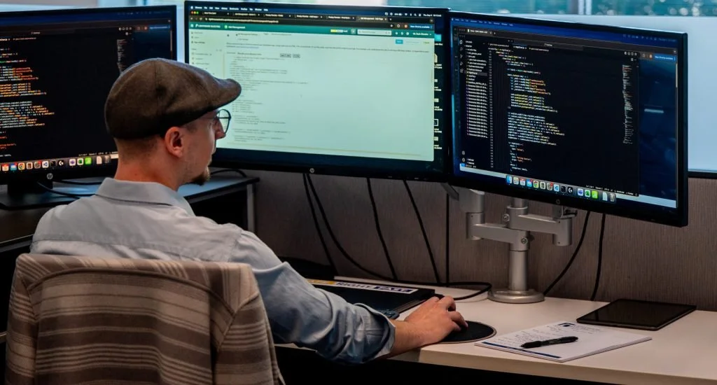 Person working at a desk with three monitors displaying code and documents, wearing glasses and a flat cap.