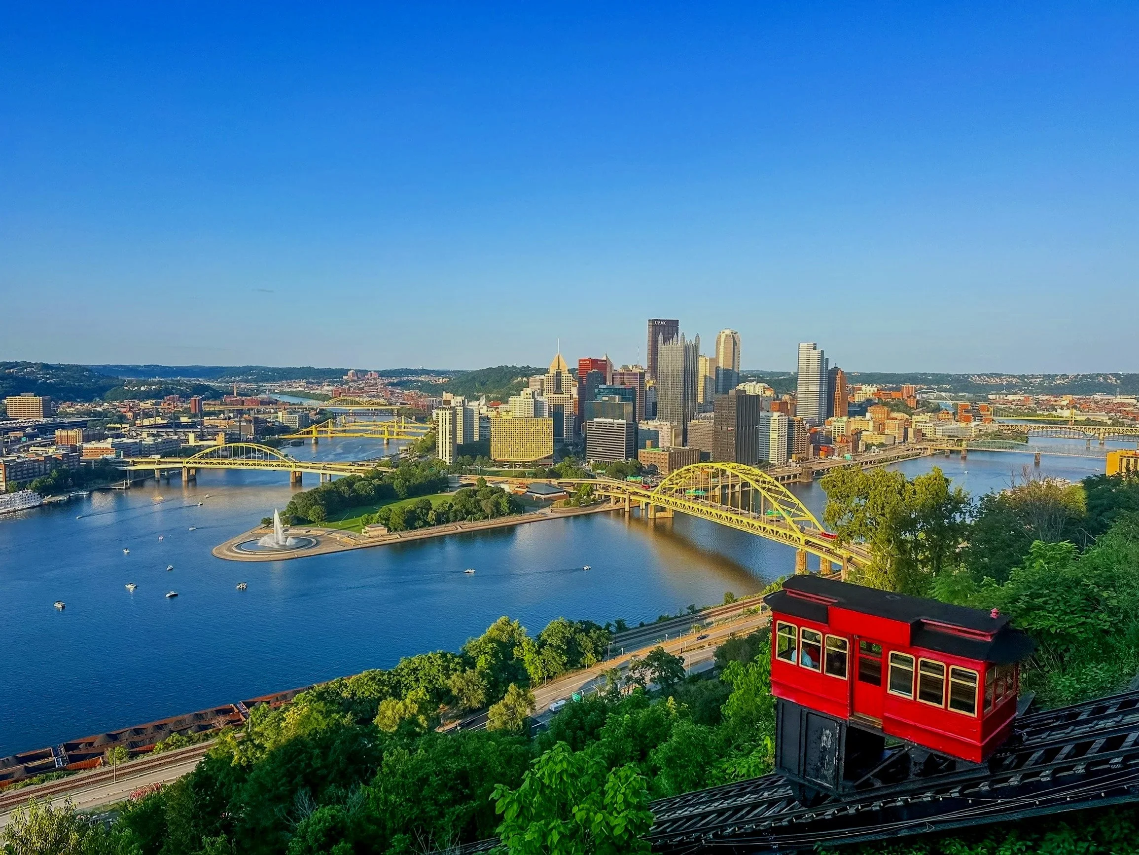 Aerial view of Pittsburgh skyline with rivers, bridges, and a red incline lift car in the foreground on a sunny day.