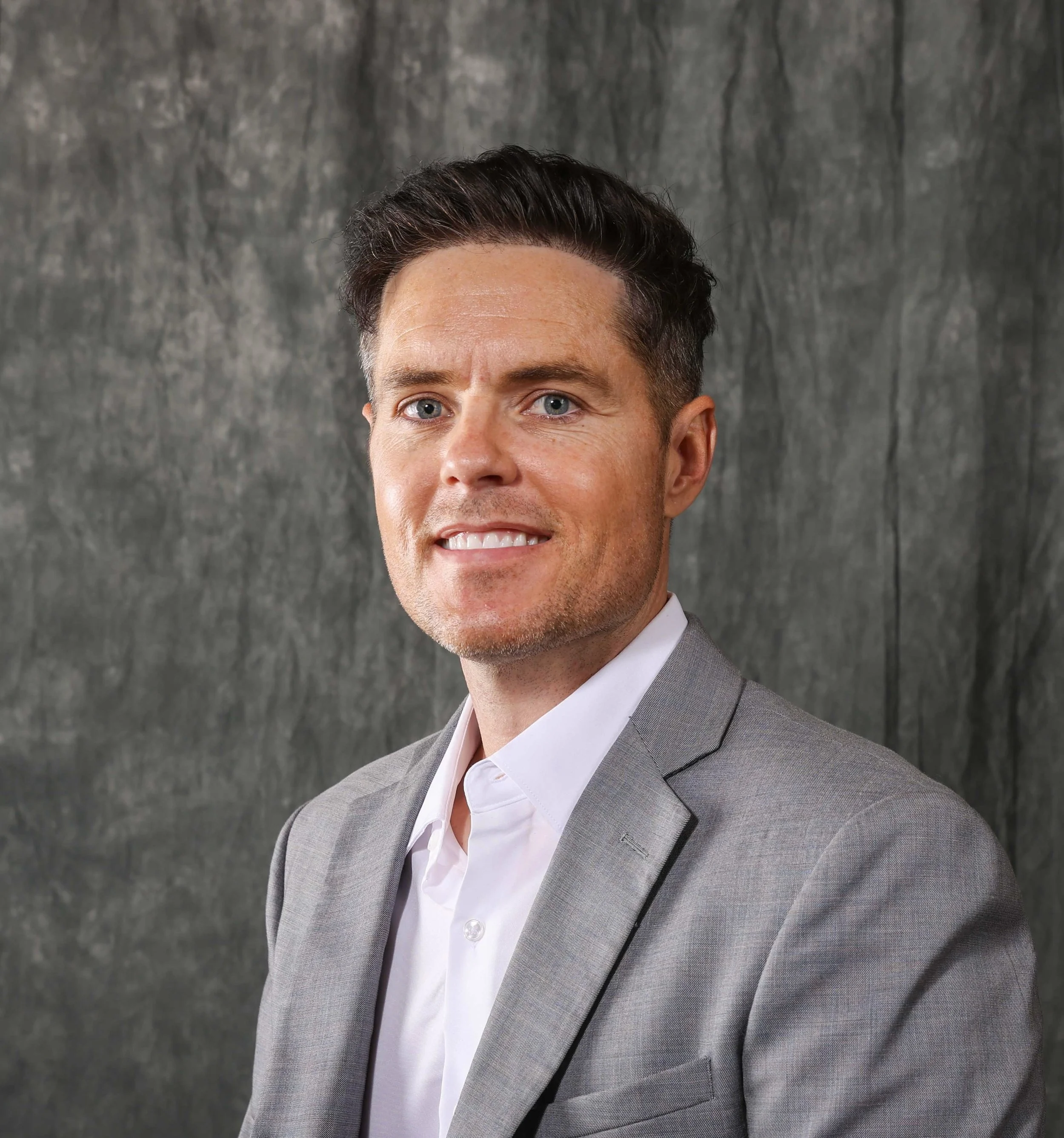 A man with short dark hair, wearing a light gray blazer and white shirt, smiling in front of a dark textured background. A photo of Nathaniel Britt Hodge. Founder of Atlas Health Systems in Lubbock, Texas.