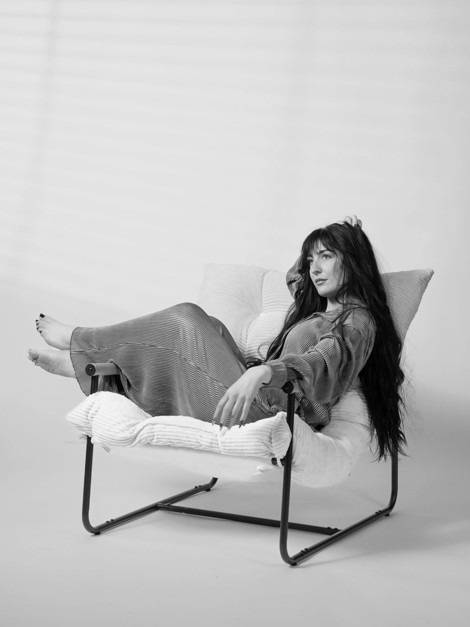 A woman with long hair reclining on a cushioned chair with a metal frame, looking thoughtfully to the side.