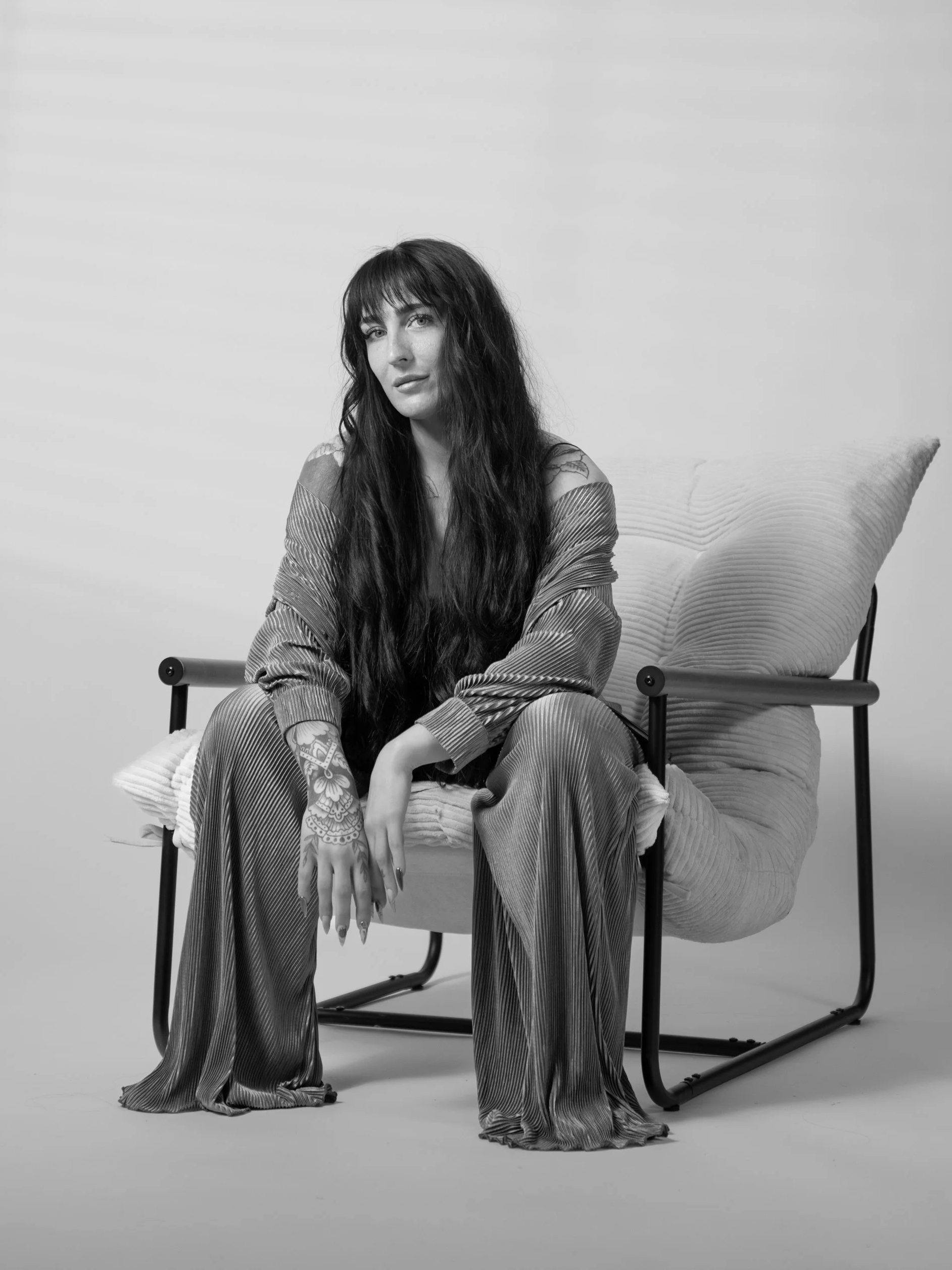 A woman with long dark hair, tattoos on her hand and shoulder, wearing a loose, shiny, pleated outfit, sitting on a modern chair with pillows in a studio setting.