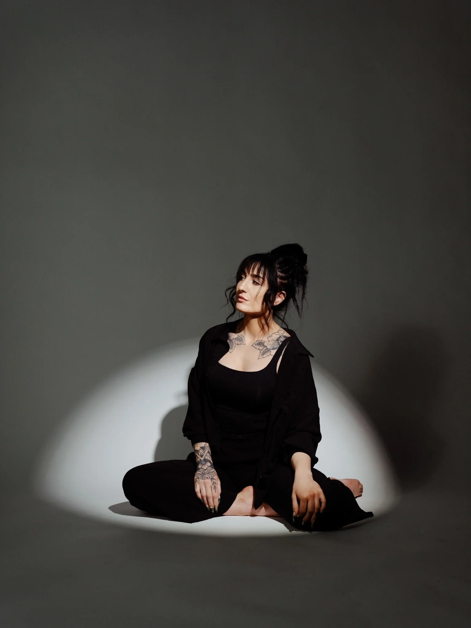 A woman with tattoos sitting cross-legged on the floor in a dark outfit, illuminated by a focused light against a dark background.