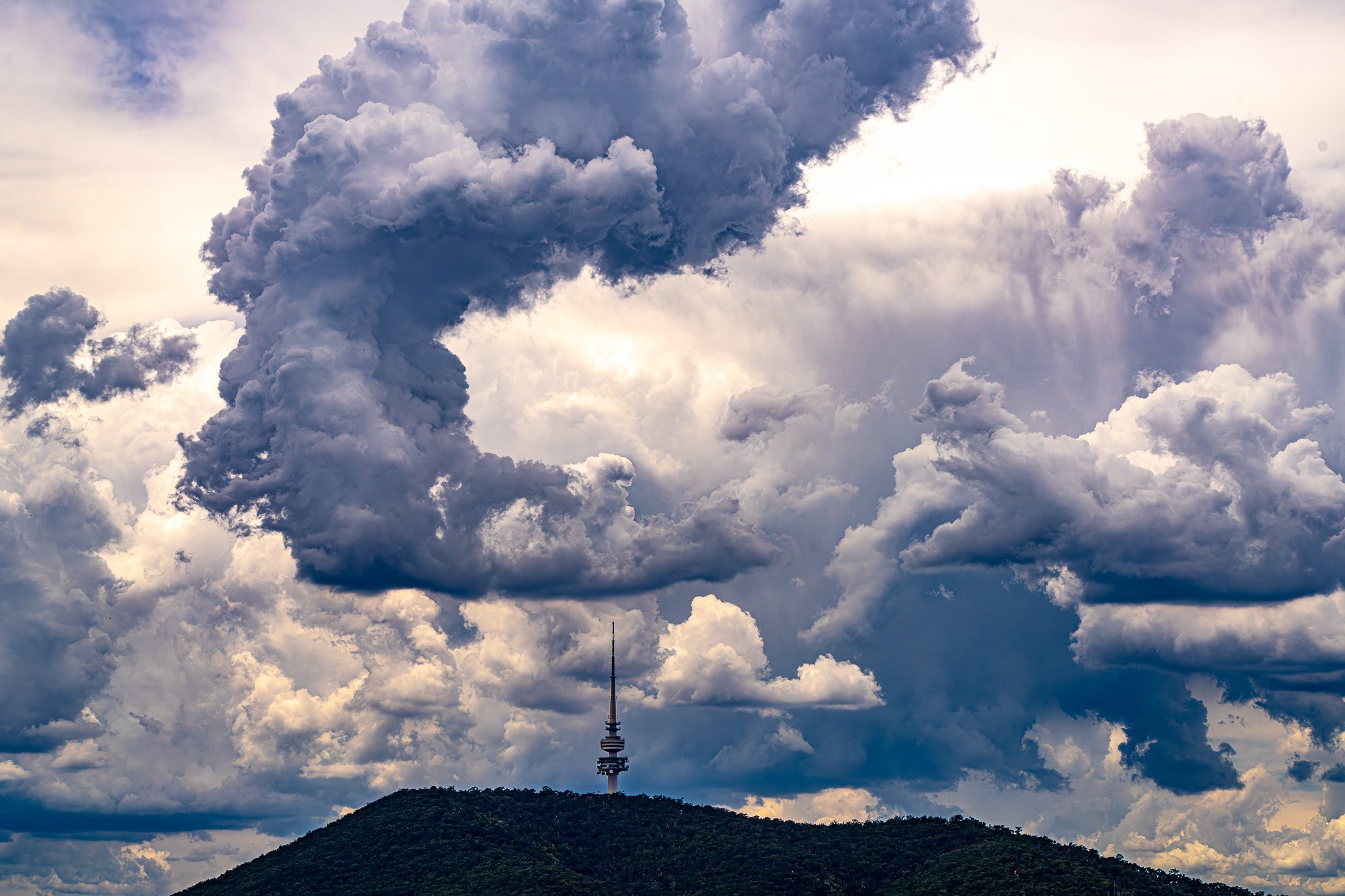 Sky filled with large gray, white, and dark clouds over a mountain with a tall communications tower (Black Mountain Tower in Canberra).