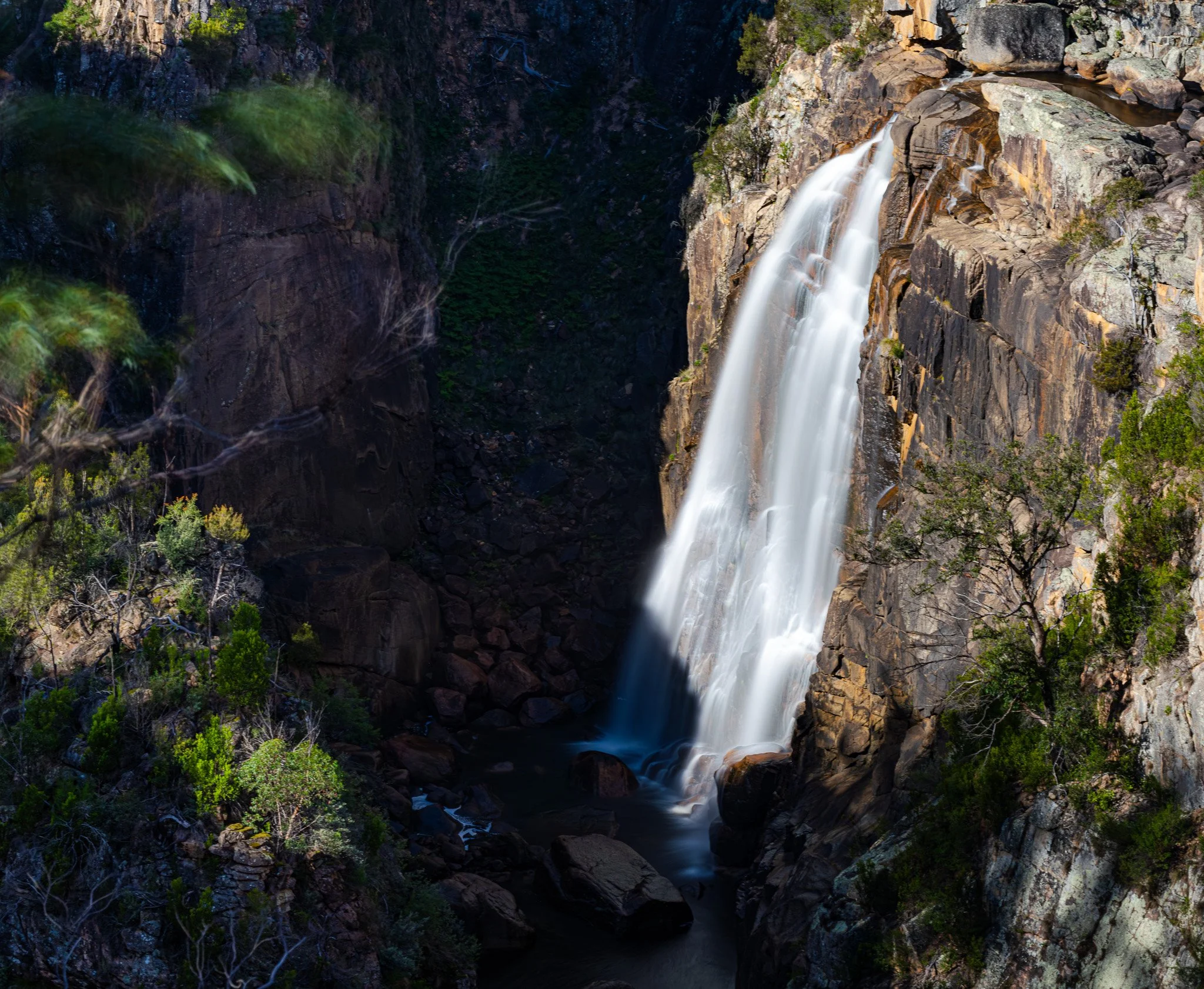 A tall waterfall cascading down a rocky cliff surrounded by trees and greenery in a natural mountainous landscape.