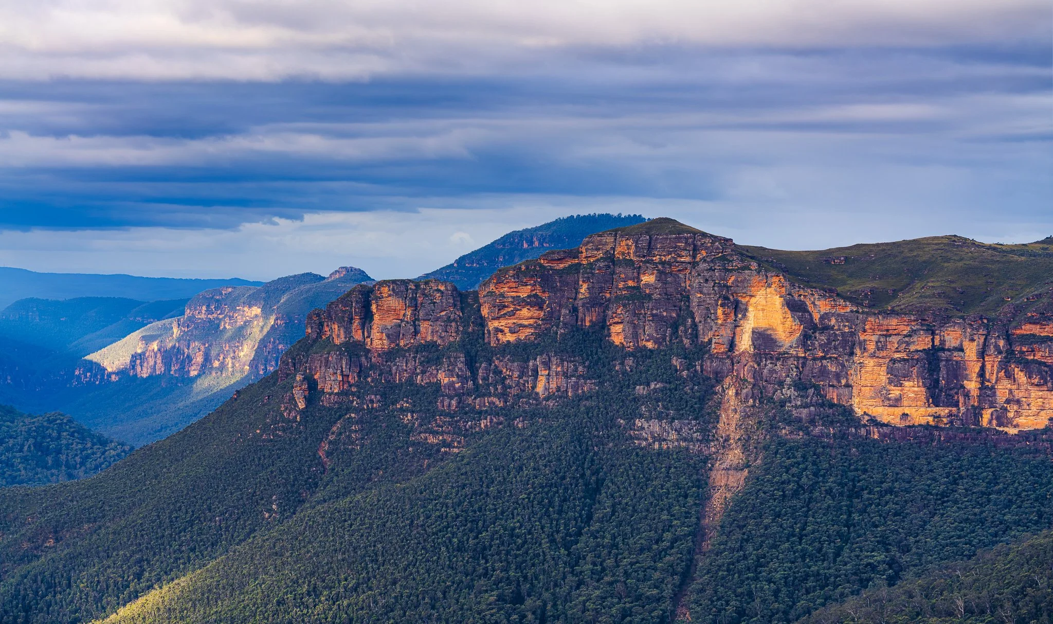 View of cliffs at the Blue Mountains, New South Wales, Australia, with green forested slopes and layered rock formations under a cloudy sky.