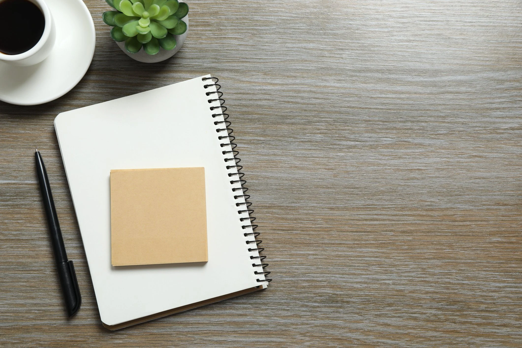 A wooden desk with a notebook, a black pen, a coffee cup, a small potted succulent plant, and a yellow sticky note.