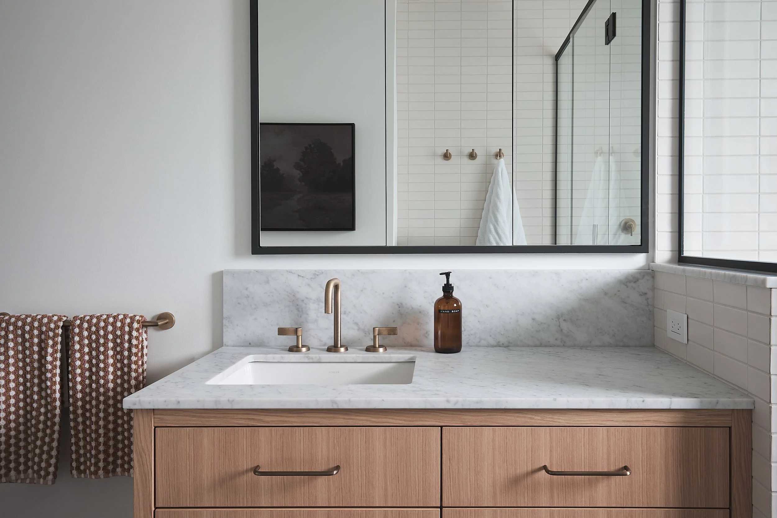 Bathroom vanity with marble countertop, oak vanity, and a large mirror. In the reflection, a shower with glass door and white tiled wall with three hooks and a towel are visible.