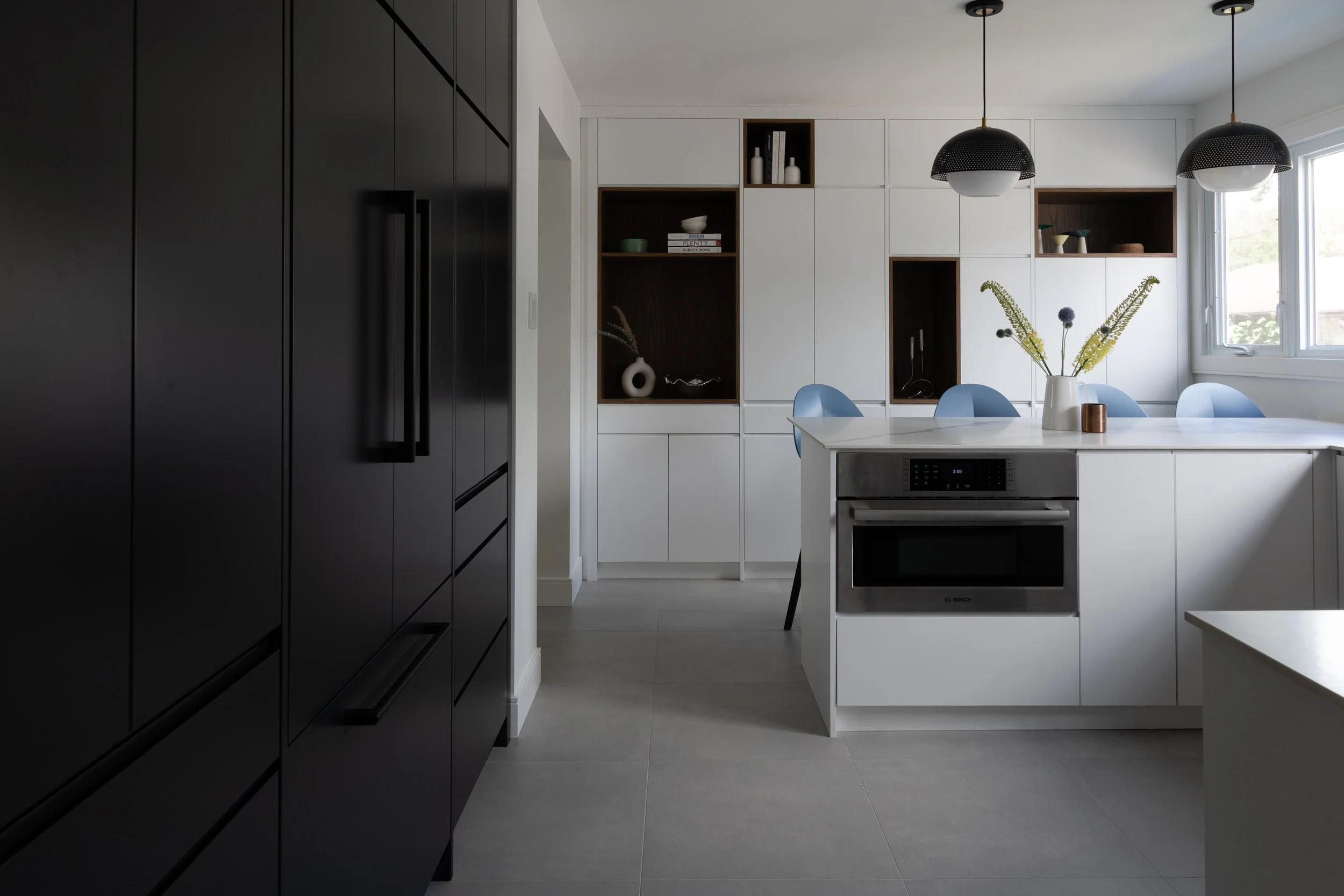 Modern kitchen with black cabinets on the left, white cabinets and a dining area with blue stools in the background, stainless steel microwave in the peninsula, and decorative vases and flowers on top near a window.