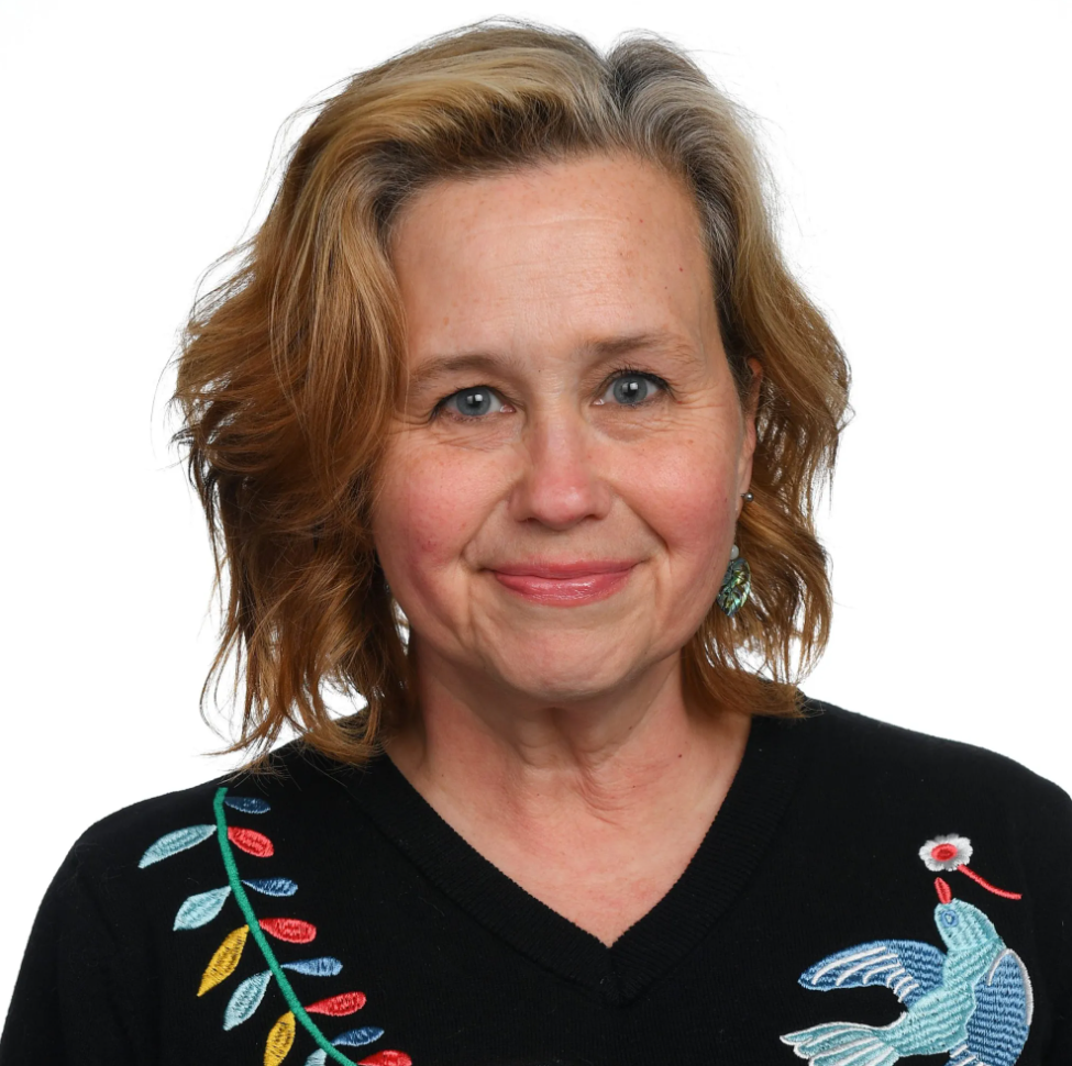 A woman with shoulder-length wavy reddish-brown hair and blue eyes, wearing a black sweater with colorful embroidered leaves and a bird design, and turquoise earrings, smiling against a white background.