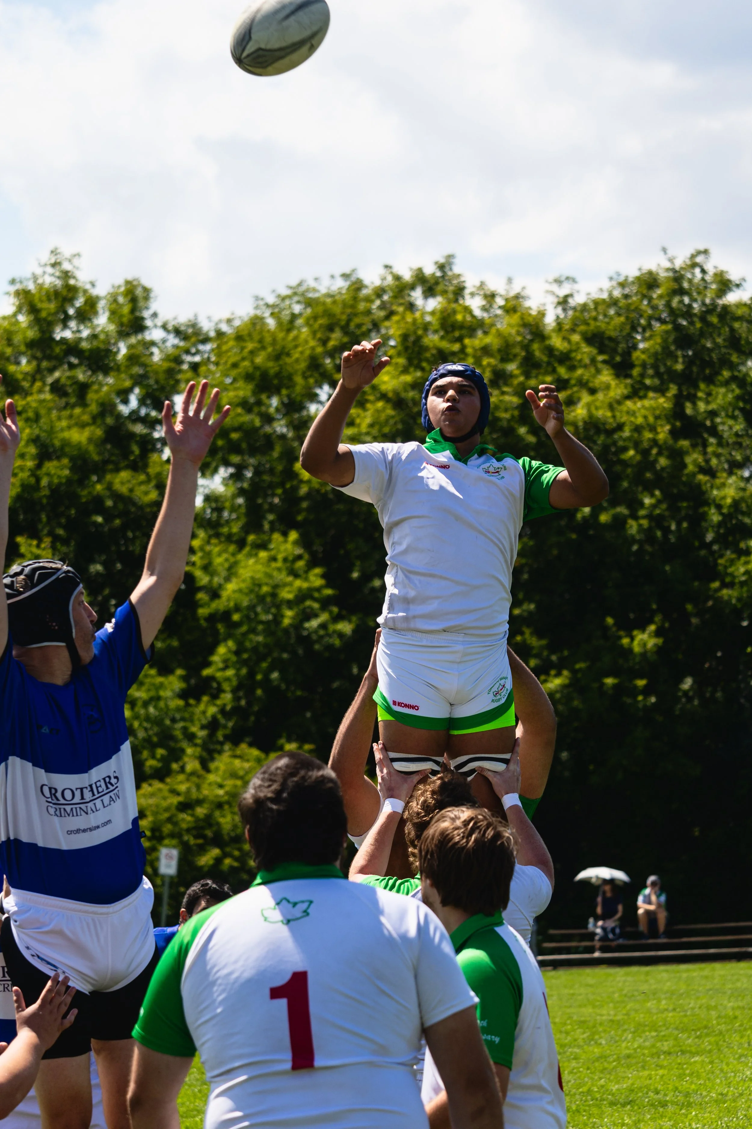 A rugby player wearing a white and green uniform is lifted by teammates during a lineout. The player is wearing a protective helmet and appears to be preparing to catch the rugby ball, which is in the air above her. The scene is outdoors in a grassy field with trees and spectators in the background.