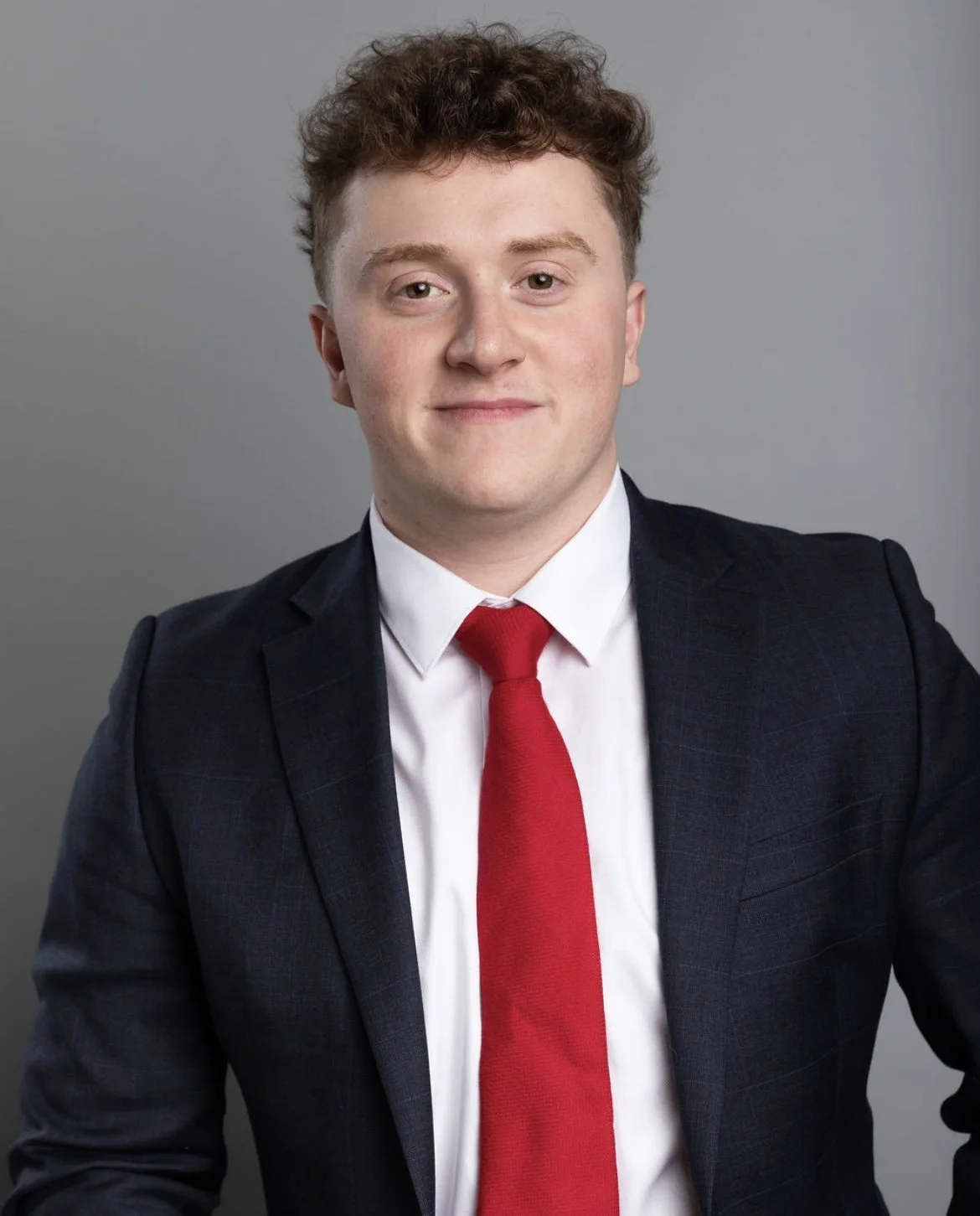 Portrait of a young man with curly hair wearing a business suit, white shirt, and red tie against a gray background.