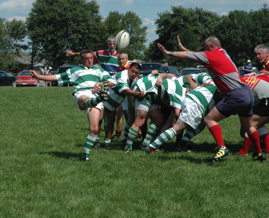 Rugby players in a scrum, wearing green and white striped jerseys, engaged in a game on a grassy field with spectators and parked cars in the background.