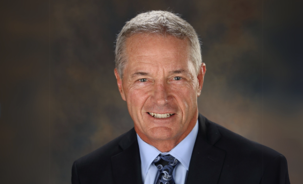 A man with gray hair wearing a black suit, light blue dress shirt, and a paisley tie, smiling against a dark, cloudy background.