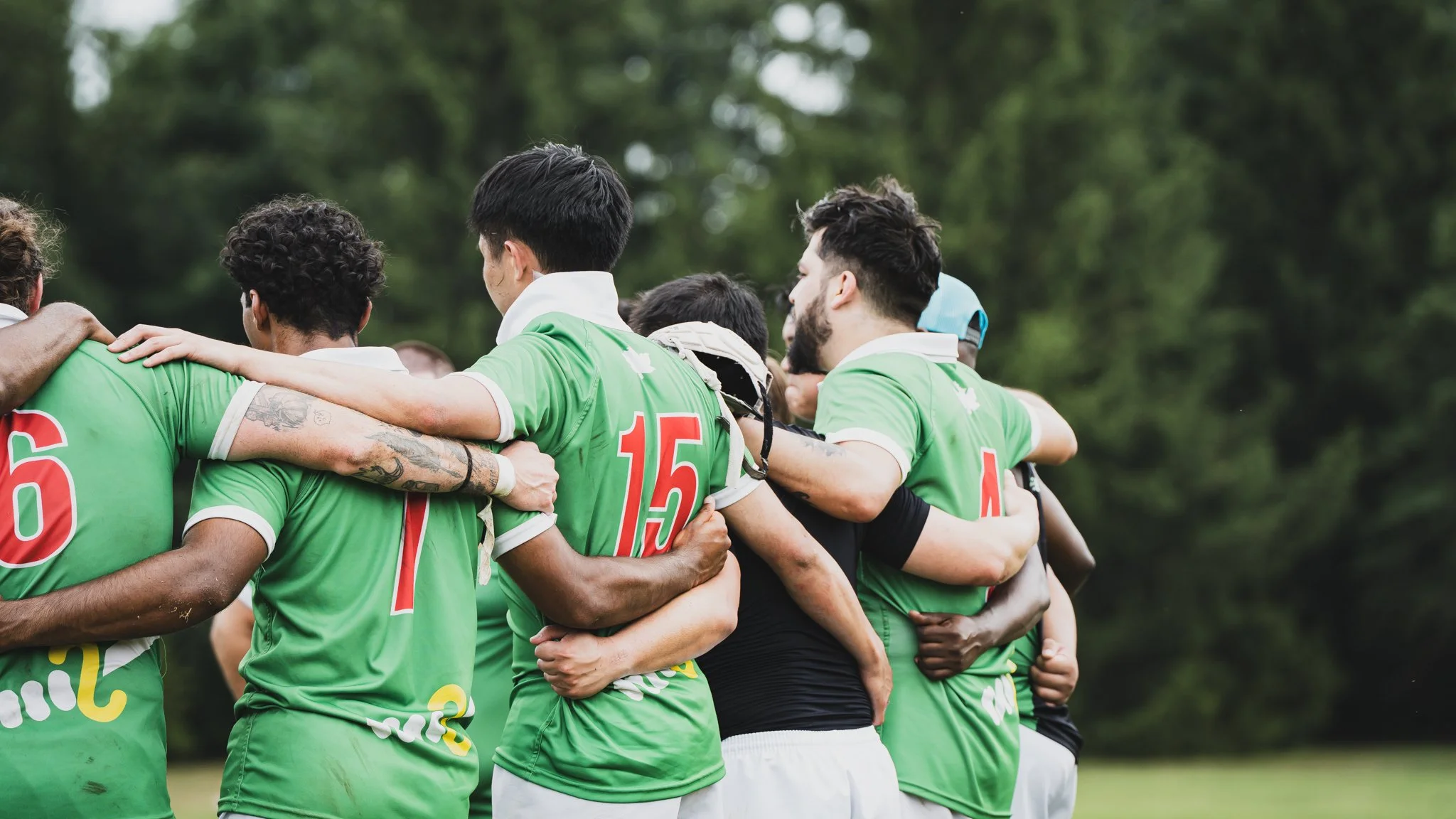 Soccer players in green jerseys huddle in a team hug on a grassy field with trees in the background.