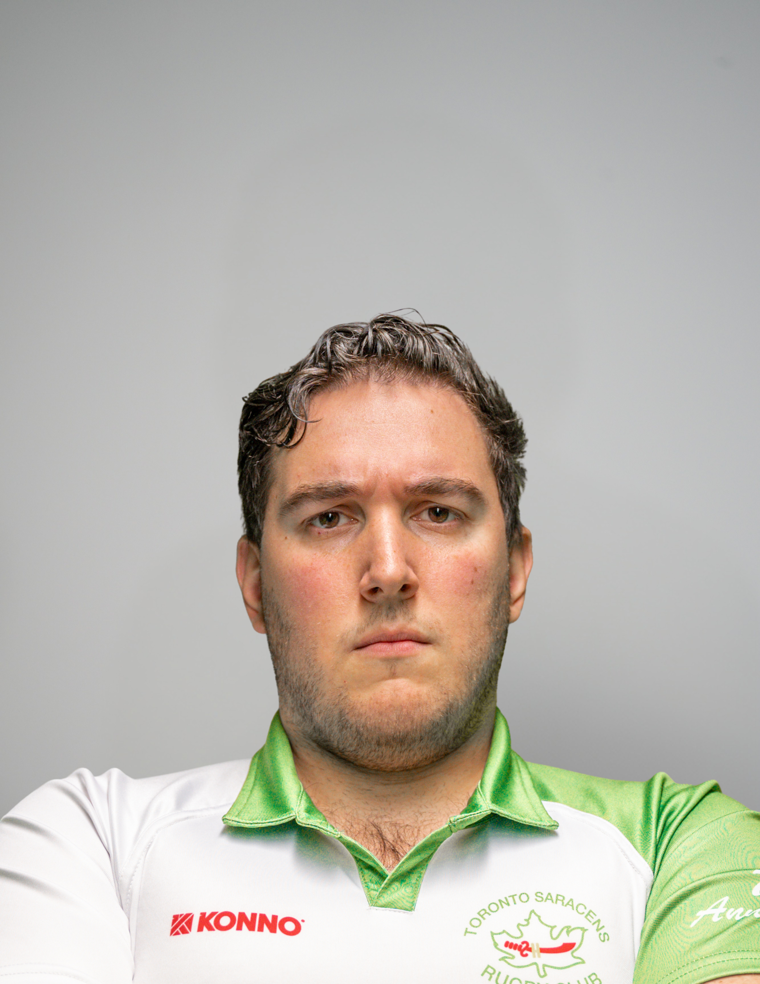 A man with short curly dark hair and light skin wearing a white and green sports jersey with logos, facing the camera against a plain gray background.