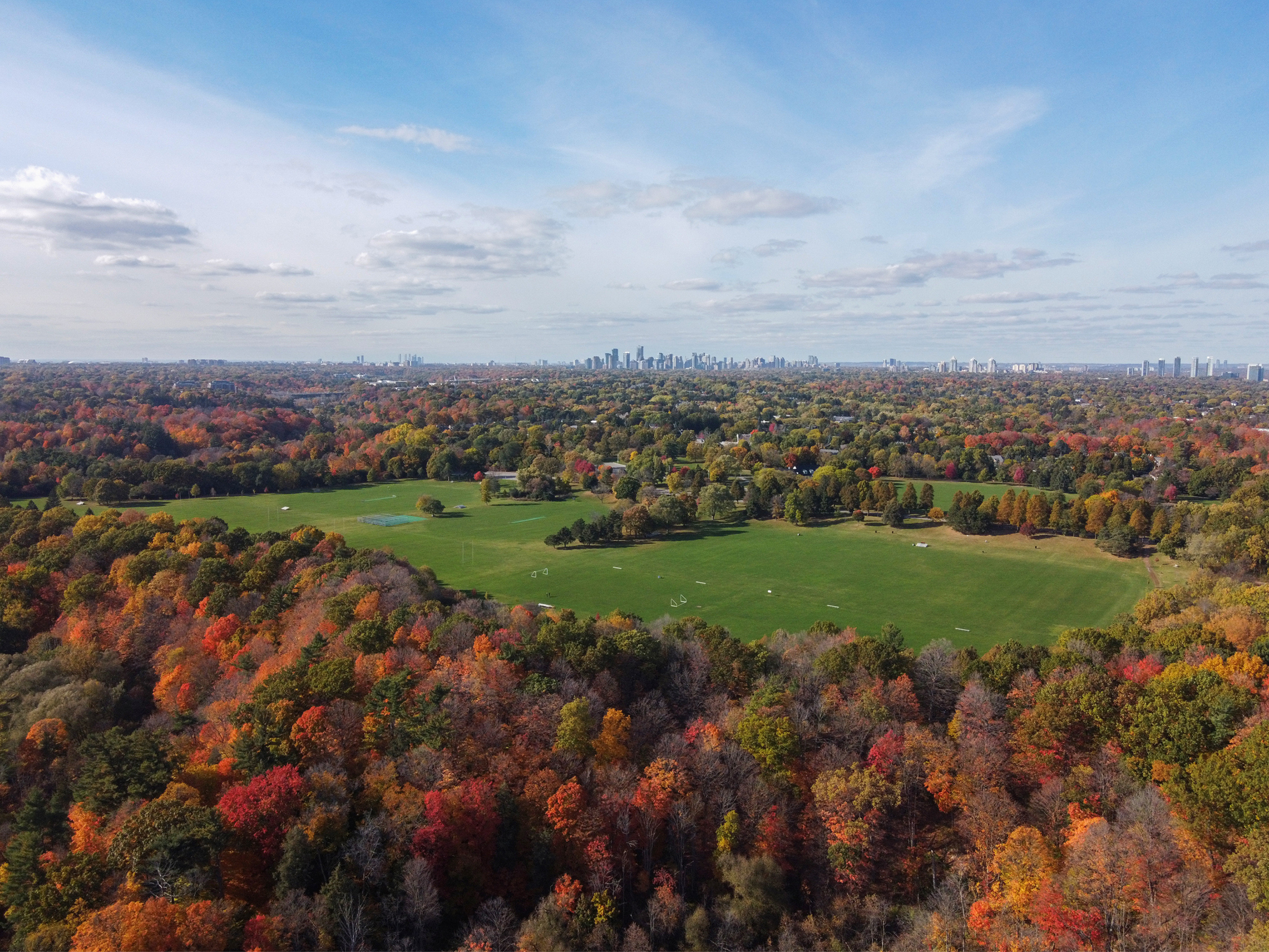 Aerial view of a city skyline with skyscrapers in the distance, surrounded by a large forest with vibrant fall foliage in shades of red, orange, yellow, and green, and a green sports field in the foreground.