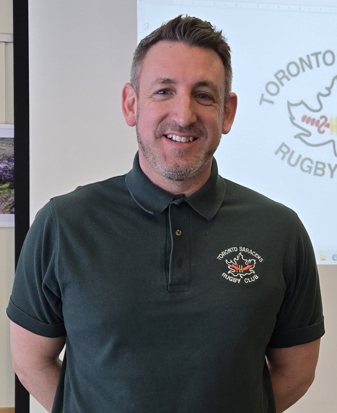 A smiling man in a dark green polo shirt with the logo of Toronto Saracens Rugby Club. Behind him, there is a projected logo of Toronto Rugby on the wall.