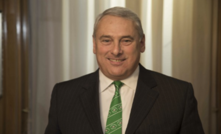 A smiling man in a suit and green tie standing indoors with curtains in the background.