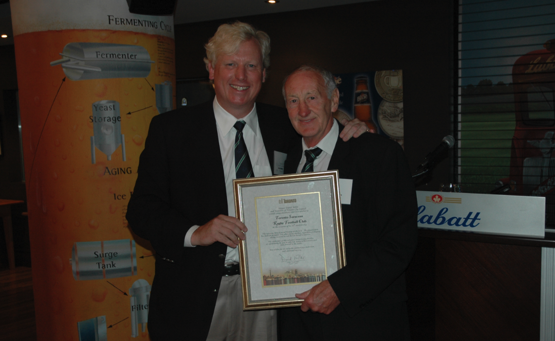 Two men dressed in black suits and striped ties stand together indoors, smiling, with one man holding a framed certificate. Behind them is a poster about beer fermentation and a window with a red truck graphic.
