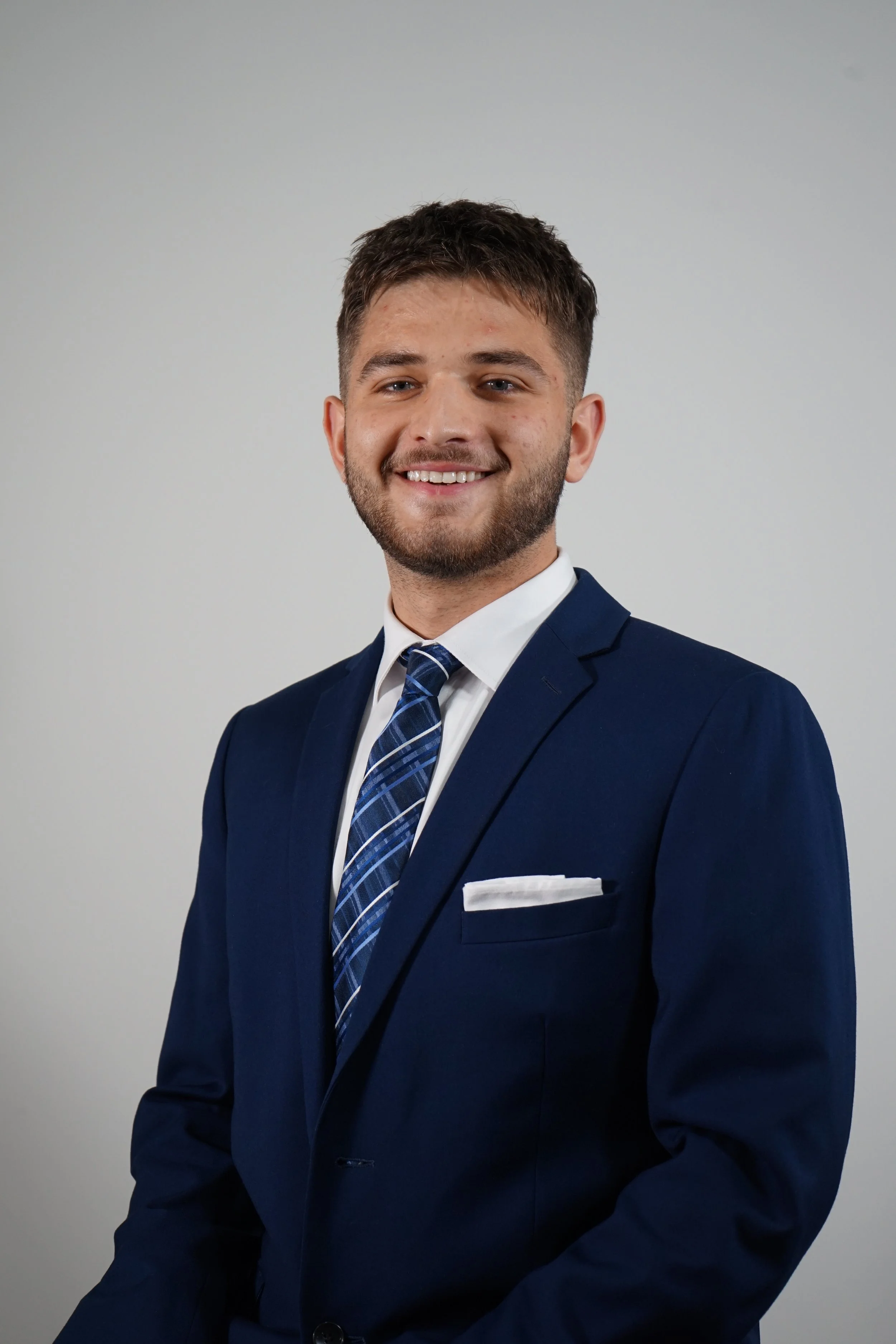 A young man in a dark blue suit, white dress shirt, and plaid tie posing in front of a plain light gray background. He has short brown hair and a beard, and he is smiling.