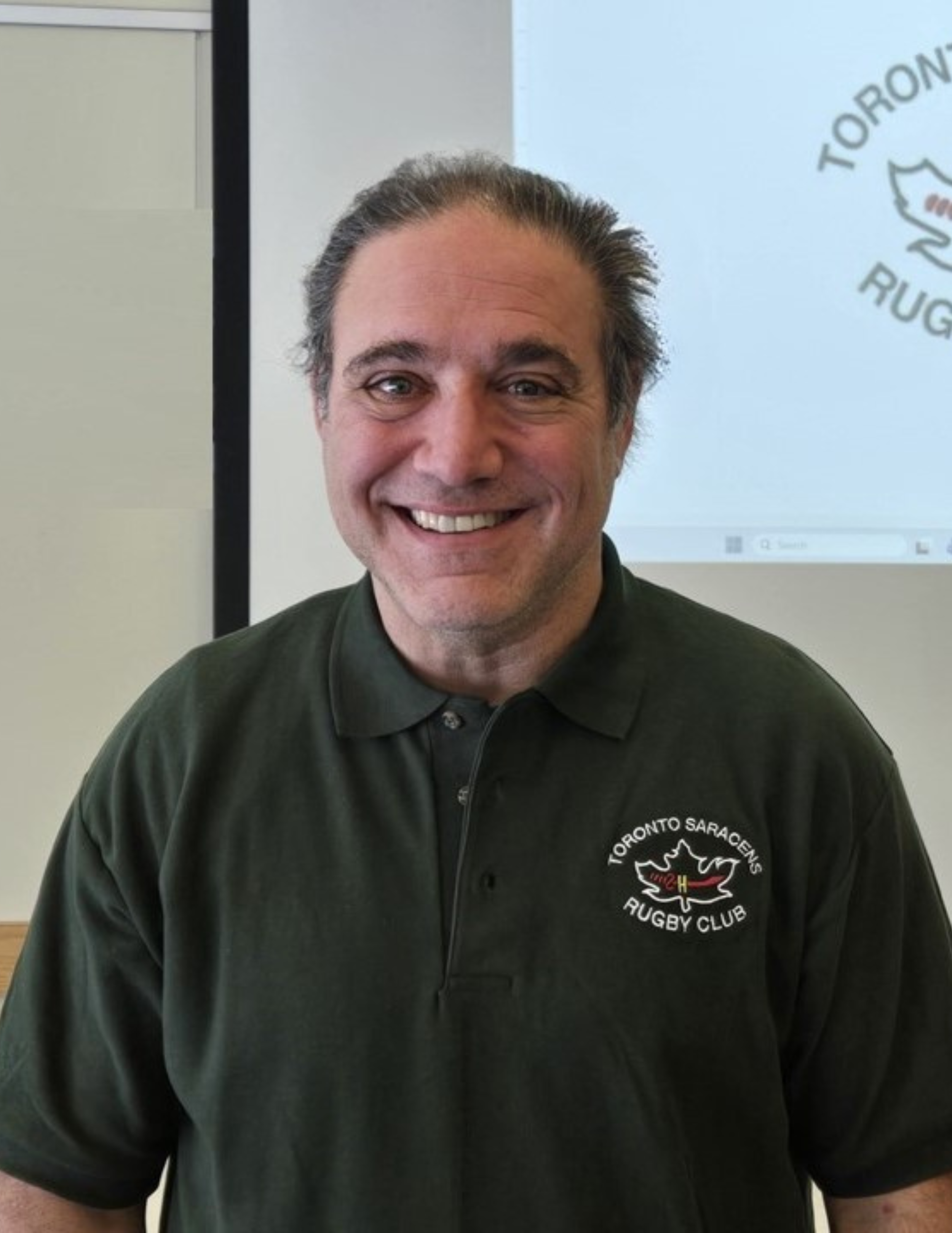 Close-up of a smiling man wearing a black collared shirt with a Toronto Saracens Rugby Club logo on the left chest, standing in front of a screen displaying a logo.