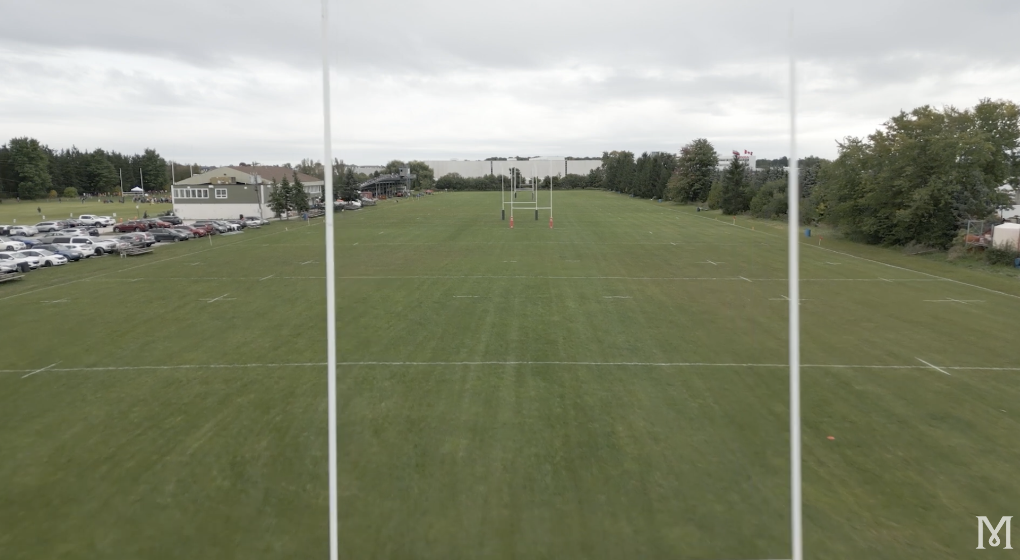View of a rugby or football field from the stands, with goal posts in the center, parking lot on the left, trees surrounding the field, and a cloudy sky overhead.