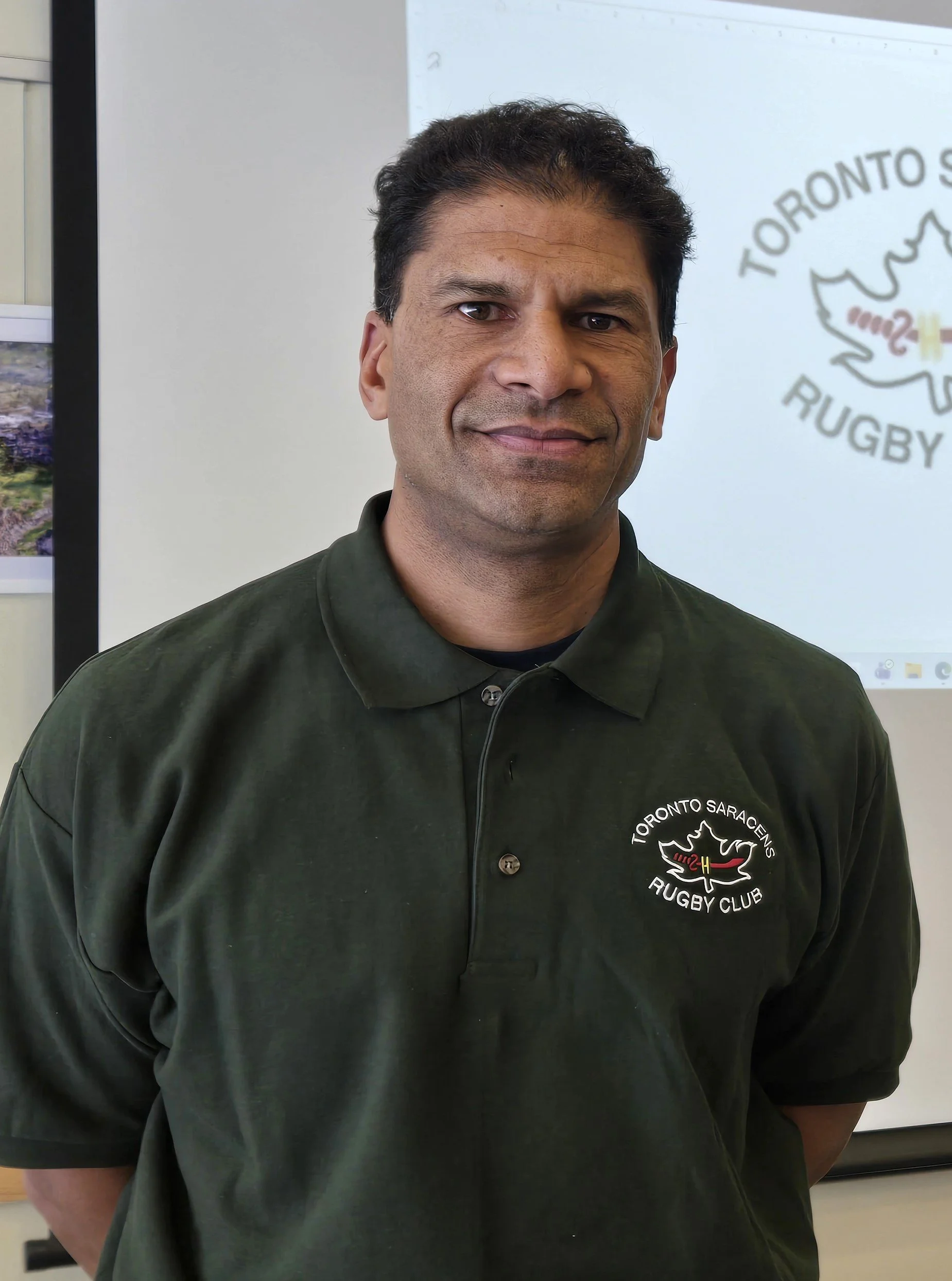 A man wearing a black polo shirt with a Toronto Saracens Rugby Club logo, standing indoors with a screen displaying the same logo in the background.