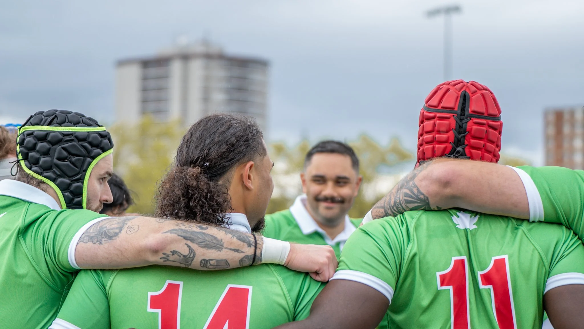 Rugby players in green jerseys huddle together with arms around each other during a game with a cityscape and cloudy sky in the background.