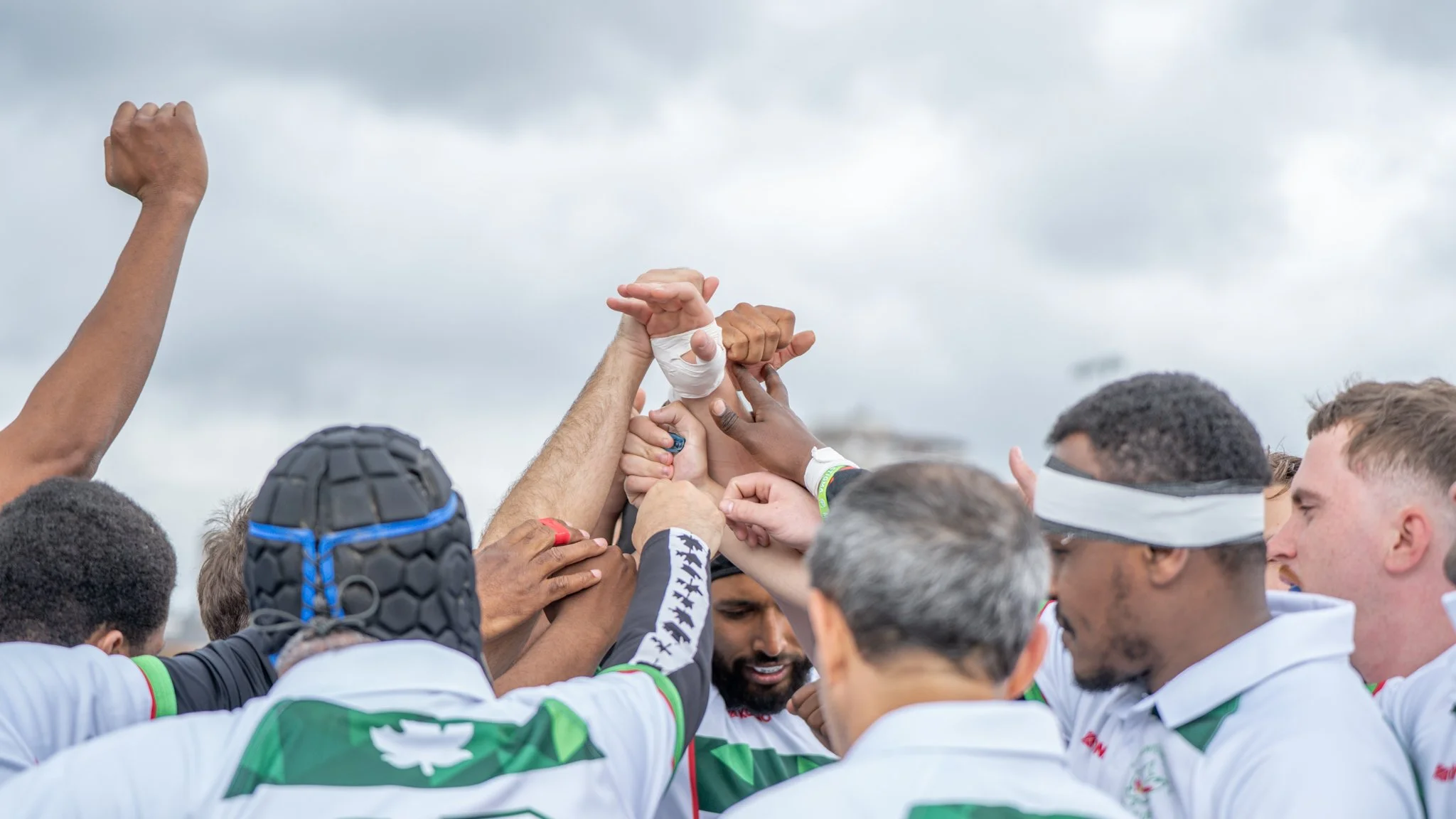 Group of athletes in a huddle raising their hands together in a gesture of unity and teamwork before a game, with cloudy sky in the background.