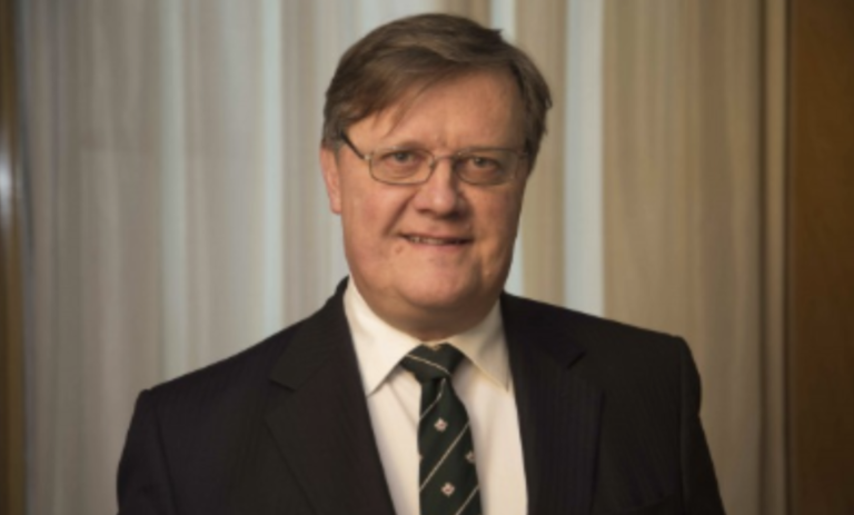 A middle-aged man with light skin and brown hair, wearing glasses, a black suit, a white shirt, and a green striped tie, standing indoors in front of beige curtains.