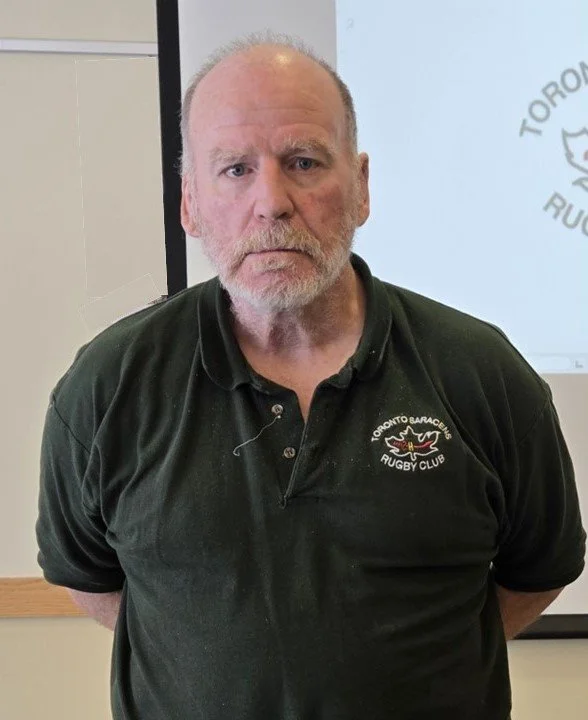 A man with a graying beard and receding hairline, wearing a black polo shirt with the Toronto Baracudas Rugby Club logo, standing indoors in front of a presentation screen.